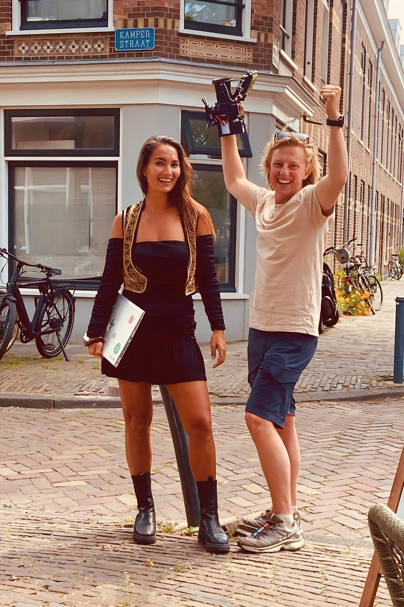 Twee lachende mensen op straat, een vrouw met lang haar en een man met blonde, winderige haar, beiden met hun armen omhoog, omringd door fietsen en een typische straatomgeving.