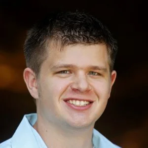 Close-up portrait of a young man with short dark hair, smiling, wearing a light blue shirt against a dark background.