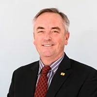 A smiling middle-aged man wearing a dark suit, white shirt, red tie, and a lapel pin, standing against a light background.