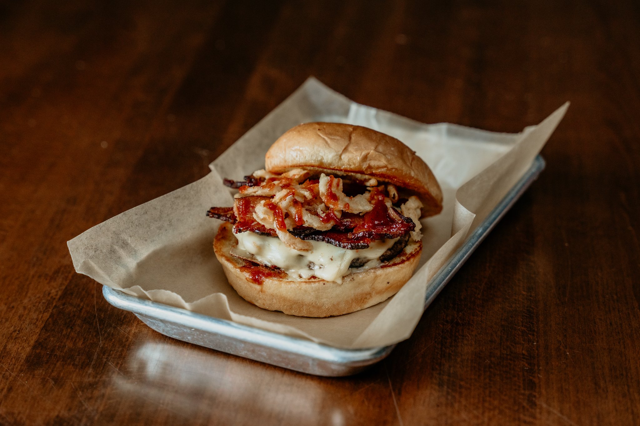 A bacon, cheese, and mushroom sandwich on a bun in a metal tray lined with parchment paper on a wooden surface.