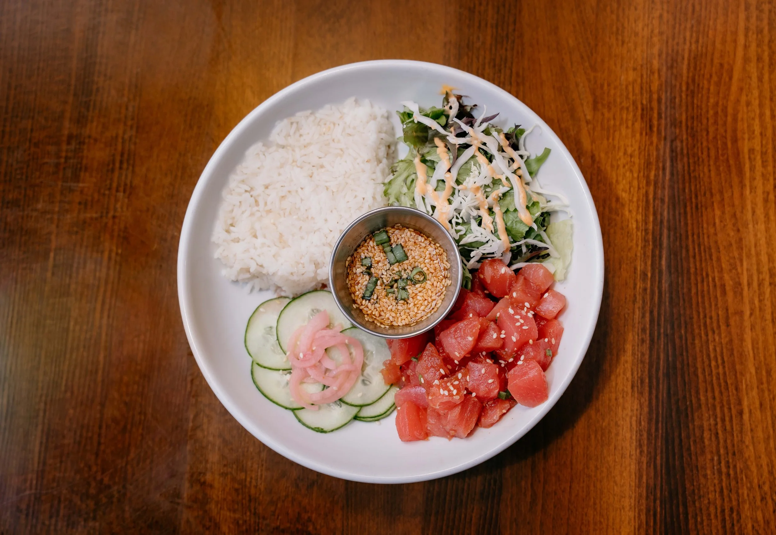 A white plate with rice, salad greens with dressing, watermelon cubes with sesame seeds, sliced cucumbers, and pickled onions, accompanied by a small container of sesame seeds garnished with chopped green onions.
