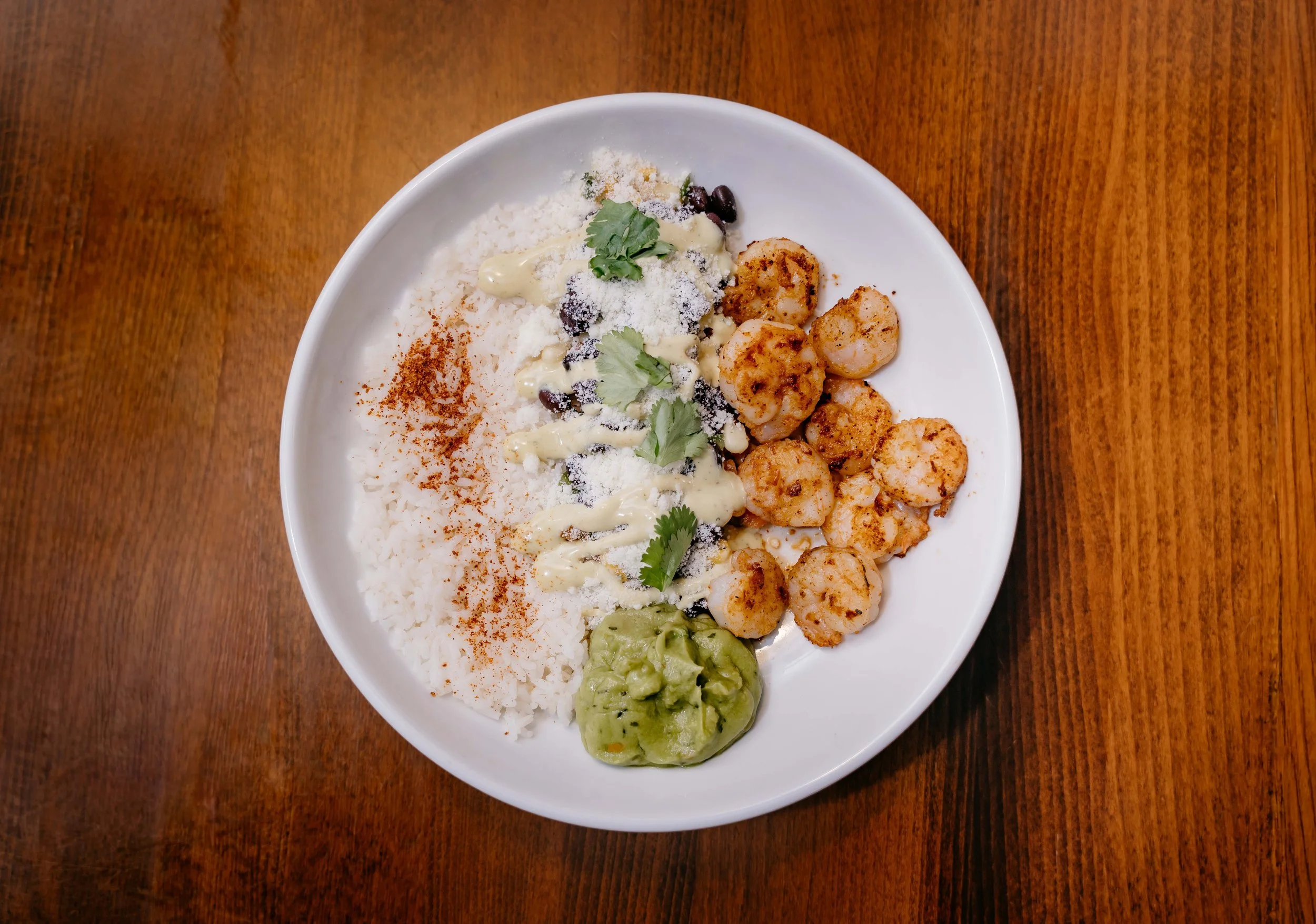 A white bowl of Mexican food with rice, grilled shrimp, avocado, black beans, shredded cheese, cilantro, and sauce on a wooden table.