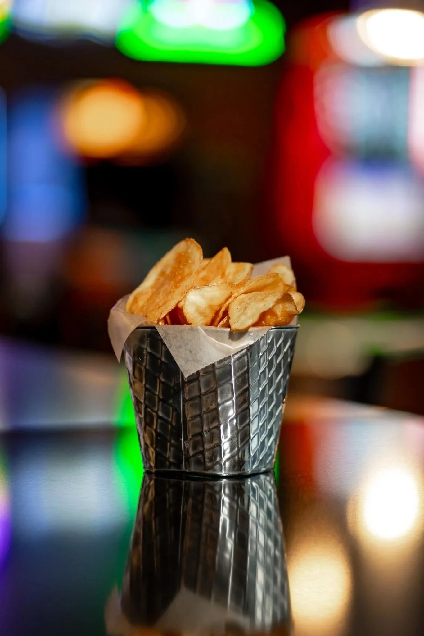 A container of potato chips on a reflective surface with colorful blurred lights in the background.