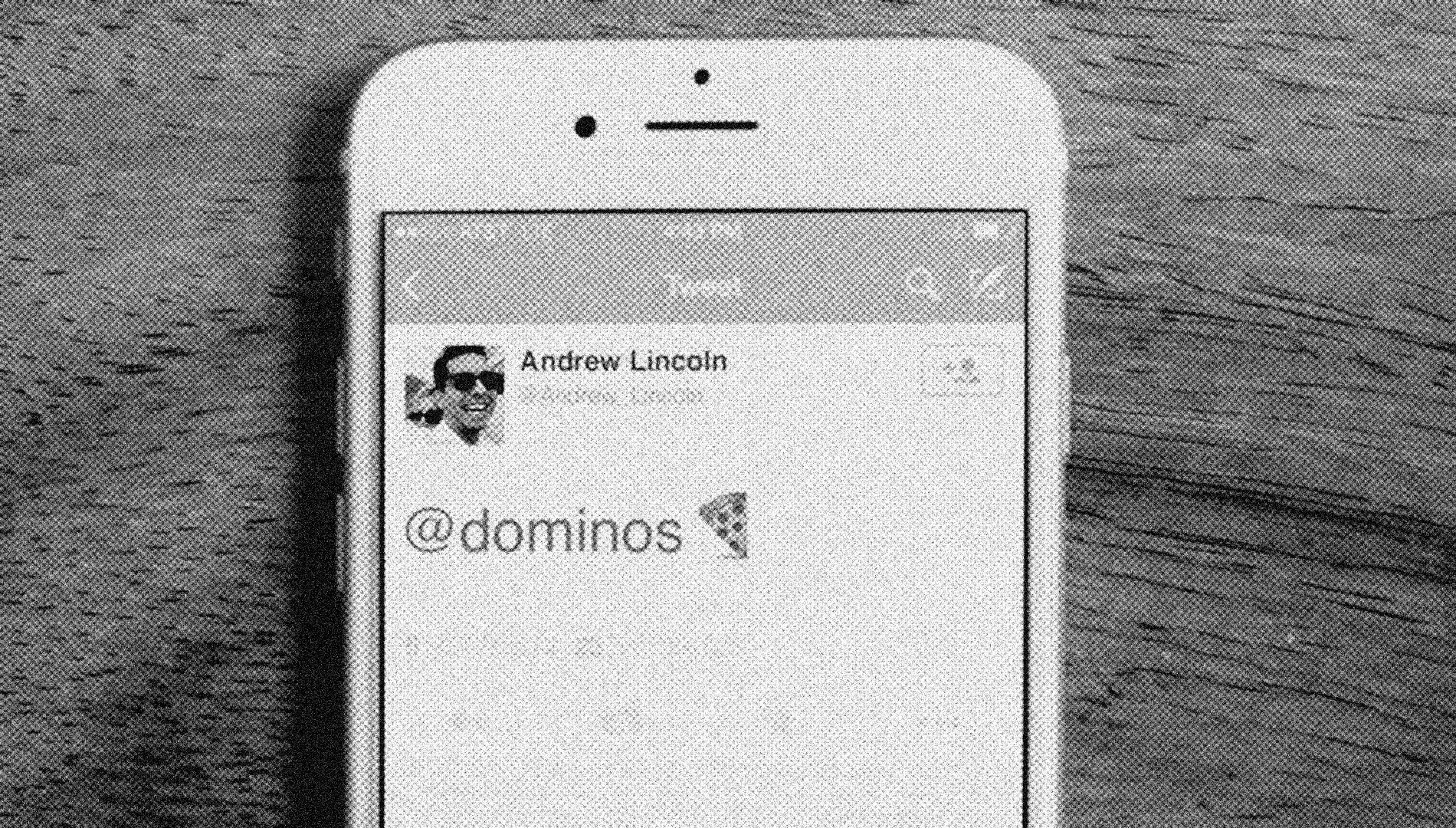 Black and white, grainy image of a smartphone screen displaying a social media post. The post shows a cartoon image of Abraham Lincoln wearing sunglasses, the name 'Andrew Lincoln,' and a caption '@dominos' with a pizza slice emoji.