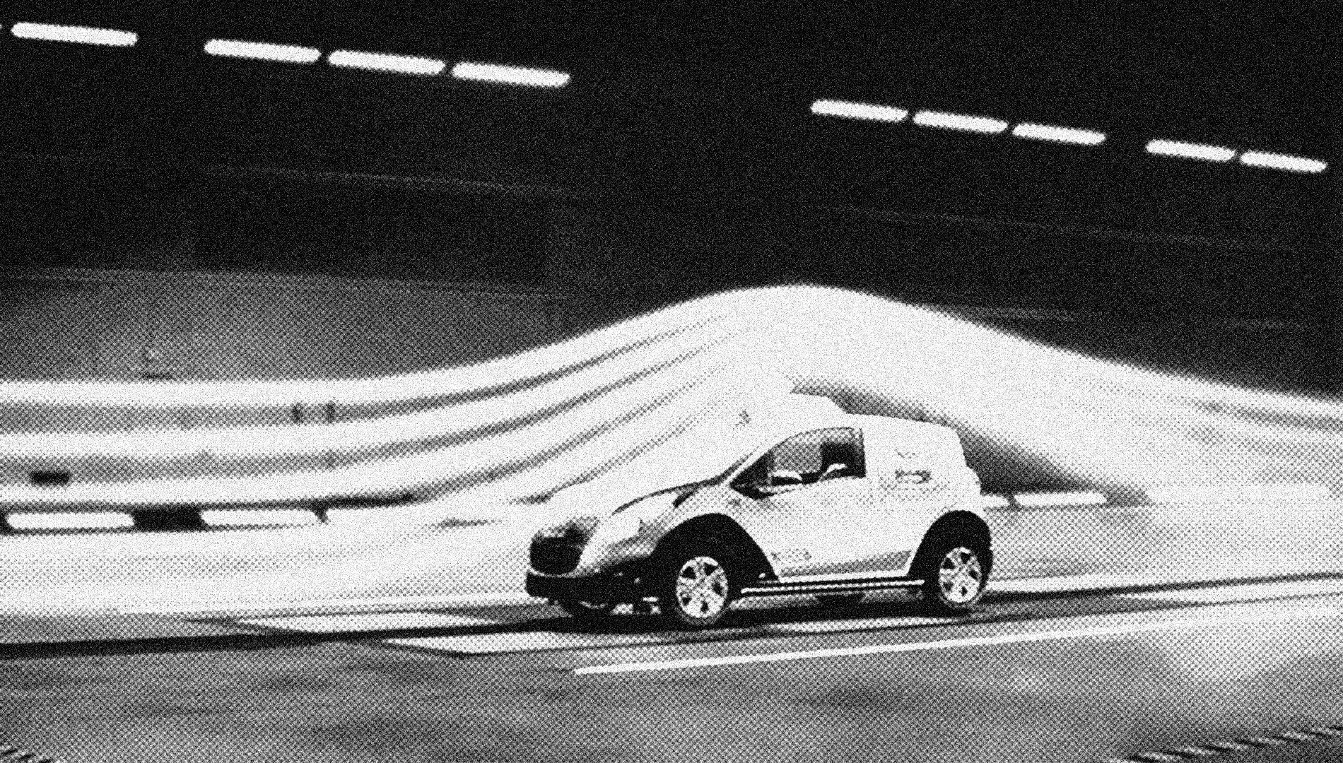 A black and white photo of a white SUV driving on a multi-lane highway at night with overpass structures overhead.