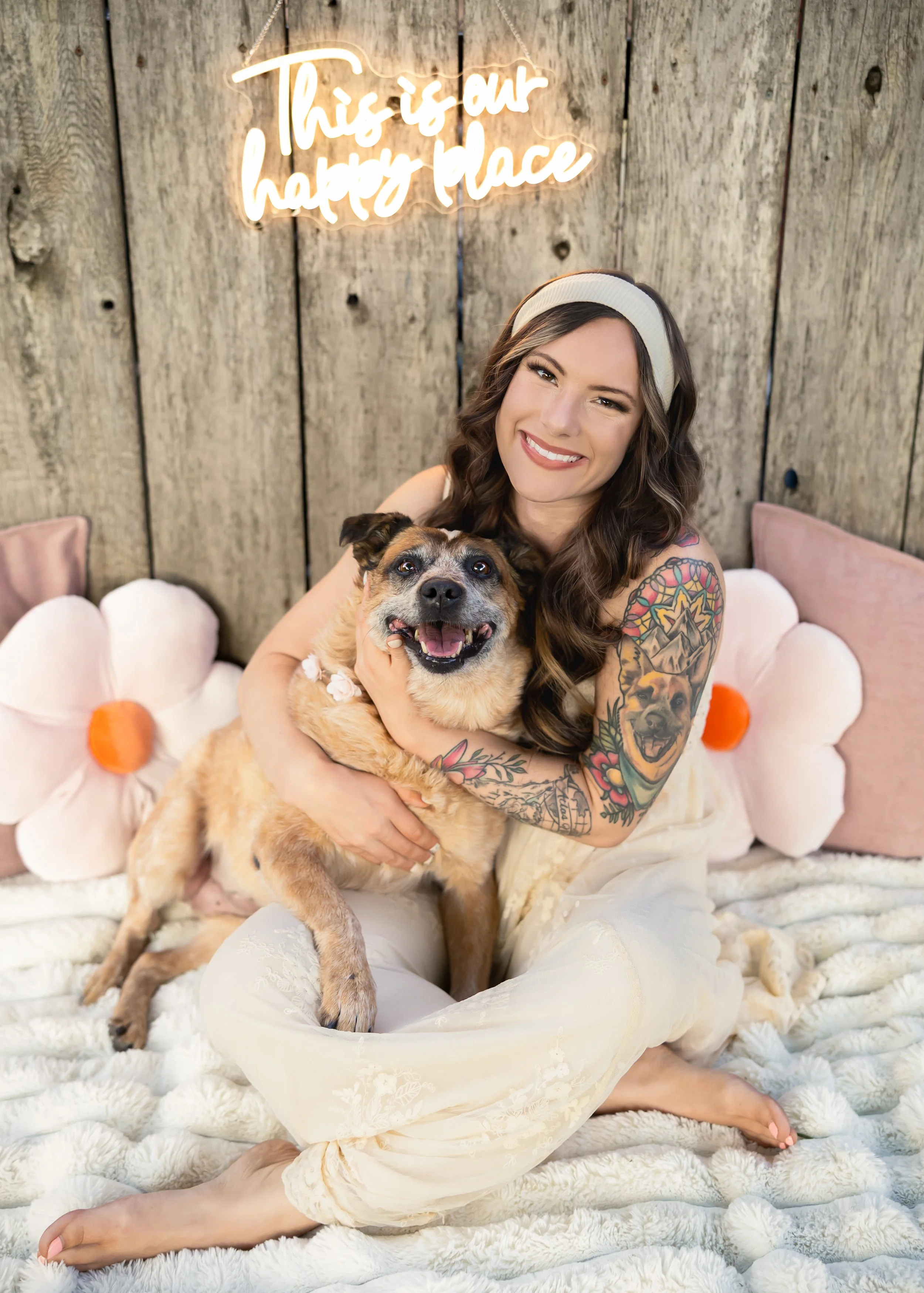 A woman with tattoos and long wavy hair, wearing a headband, sitting on a fluffy blanket, hugging a happy dog with tattoos, with a wooden wall background and a neon sign that reads 'This is our happy place'.