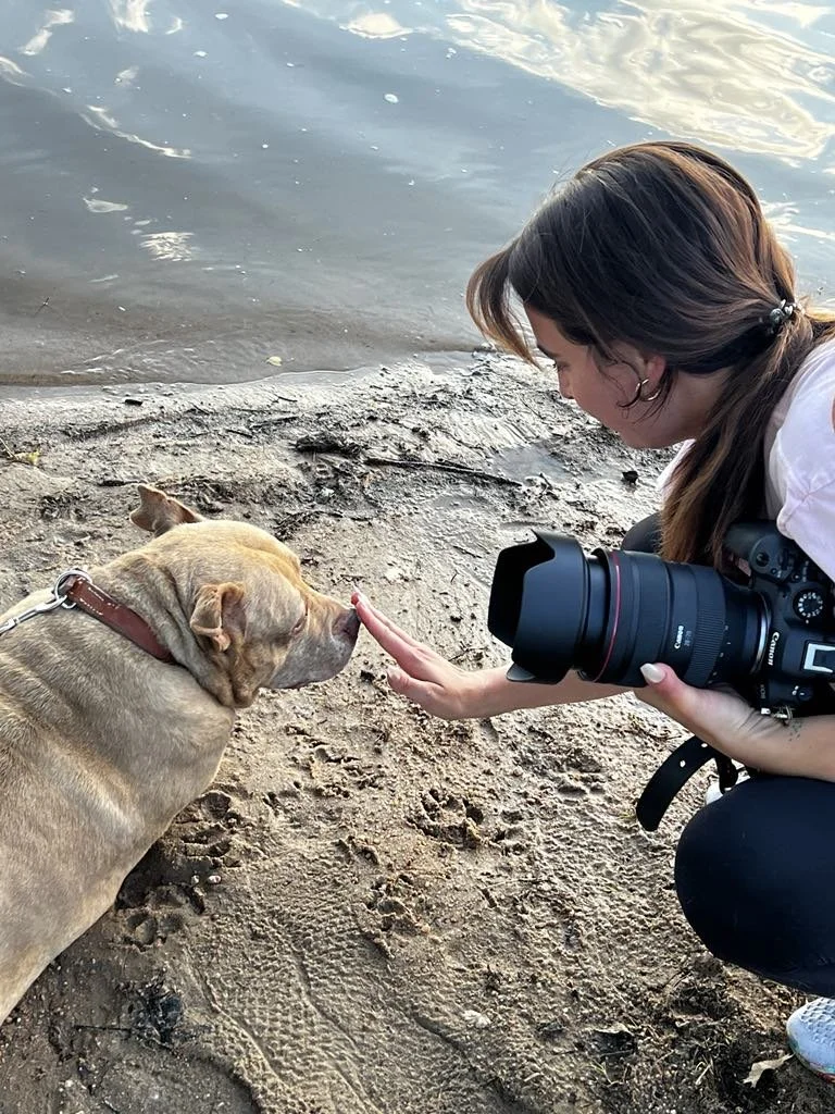 A person with brown hair, holding a camera, touching noses with a tan dog on a sandy beach by the water.