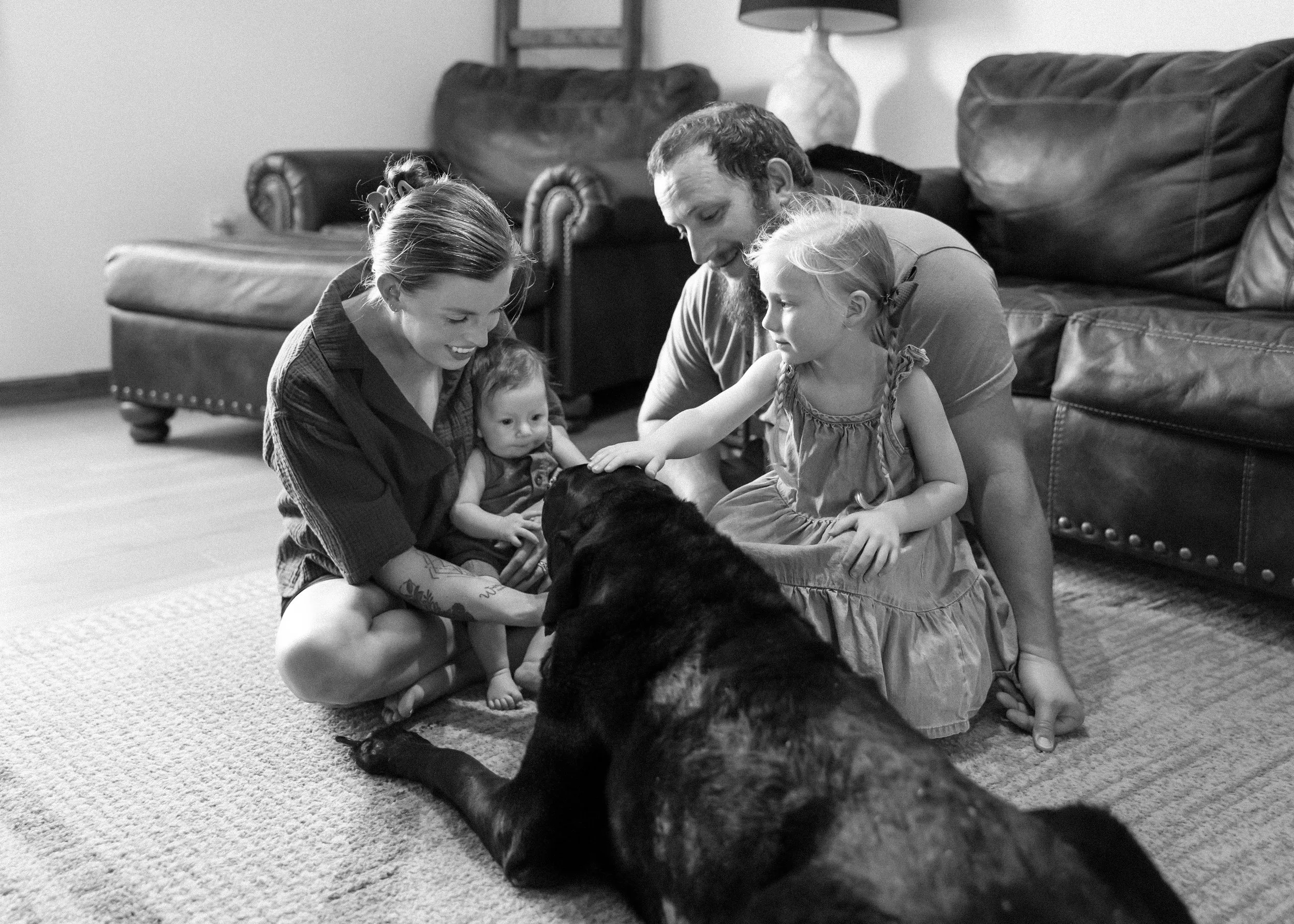 A family centered around their pet dog on a carpet in a living room, with parents and two young girls, engaging with and petting the dog.