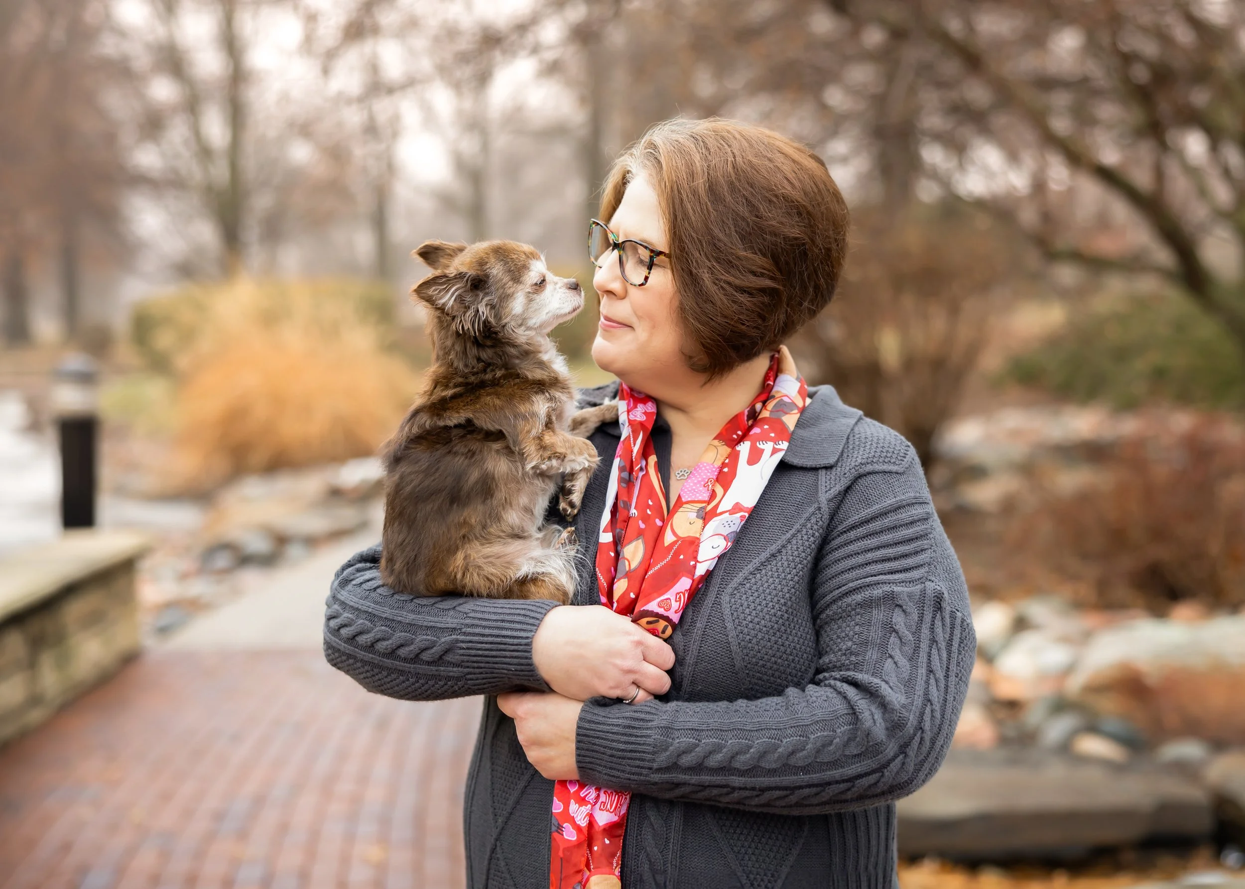 A woman holding a small brown and white dog outdoors in a park during fall, looking into the dog's face with a smile.