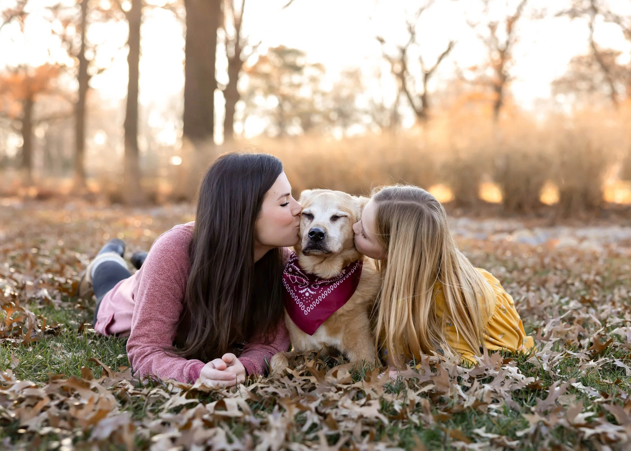 Two young girls lying on autumn leaves in a park, kissing a light-colored dog with a bandana around its neck.