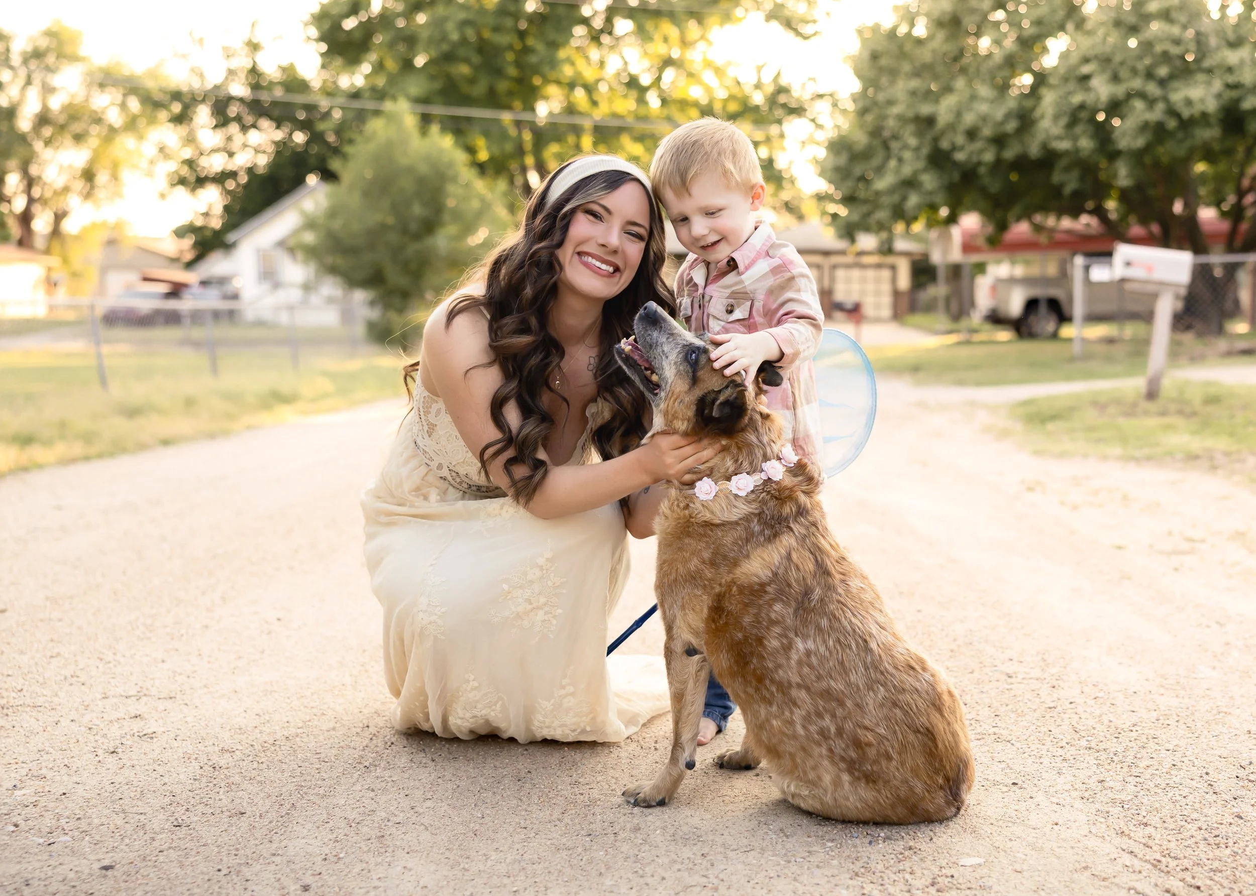 Smiling woman in a beige dress kneeling on ground, holding a dog with a flower necklace, while a young boy in a plaid shirt petting the dog, during sunset in a suburban neighborhood.