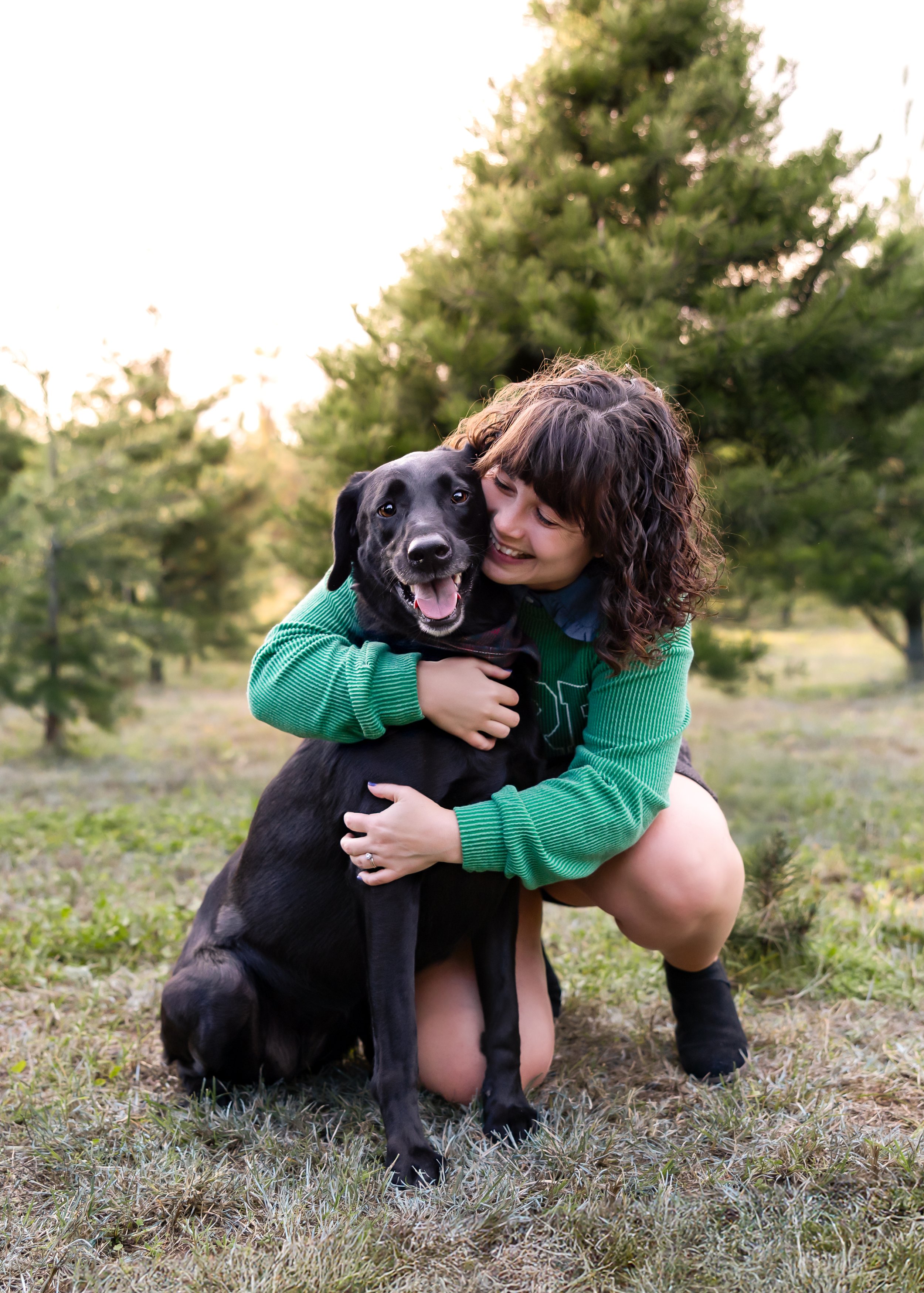 A young woman smiling and hugging a black dog while kneeling on grass in a park with trees in the background.