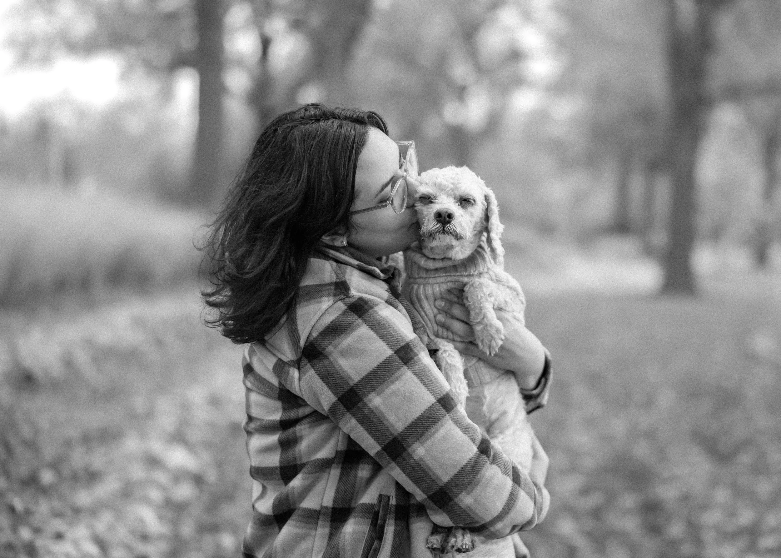 A woman with glasses and dark hair, wearing a plaid jacket, holding a small dog in a sweater, while giving it a kiss on the face in an outdoor park setting.