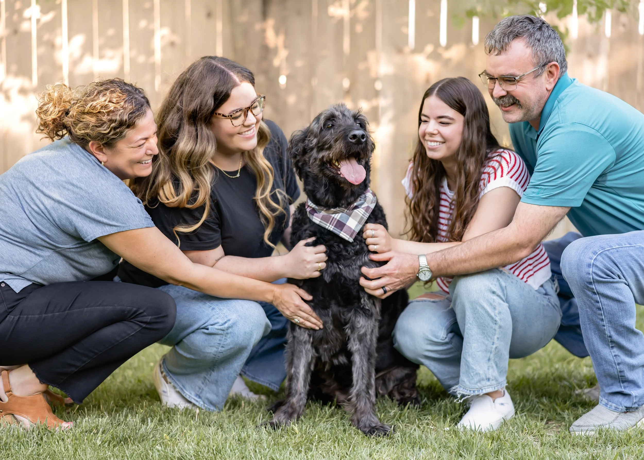 Family of five people and a black dog with a bandana outdoors in a backyard, smiling and playing.
