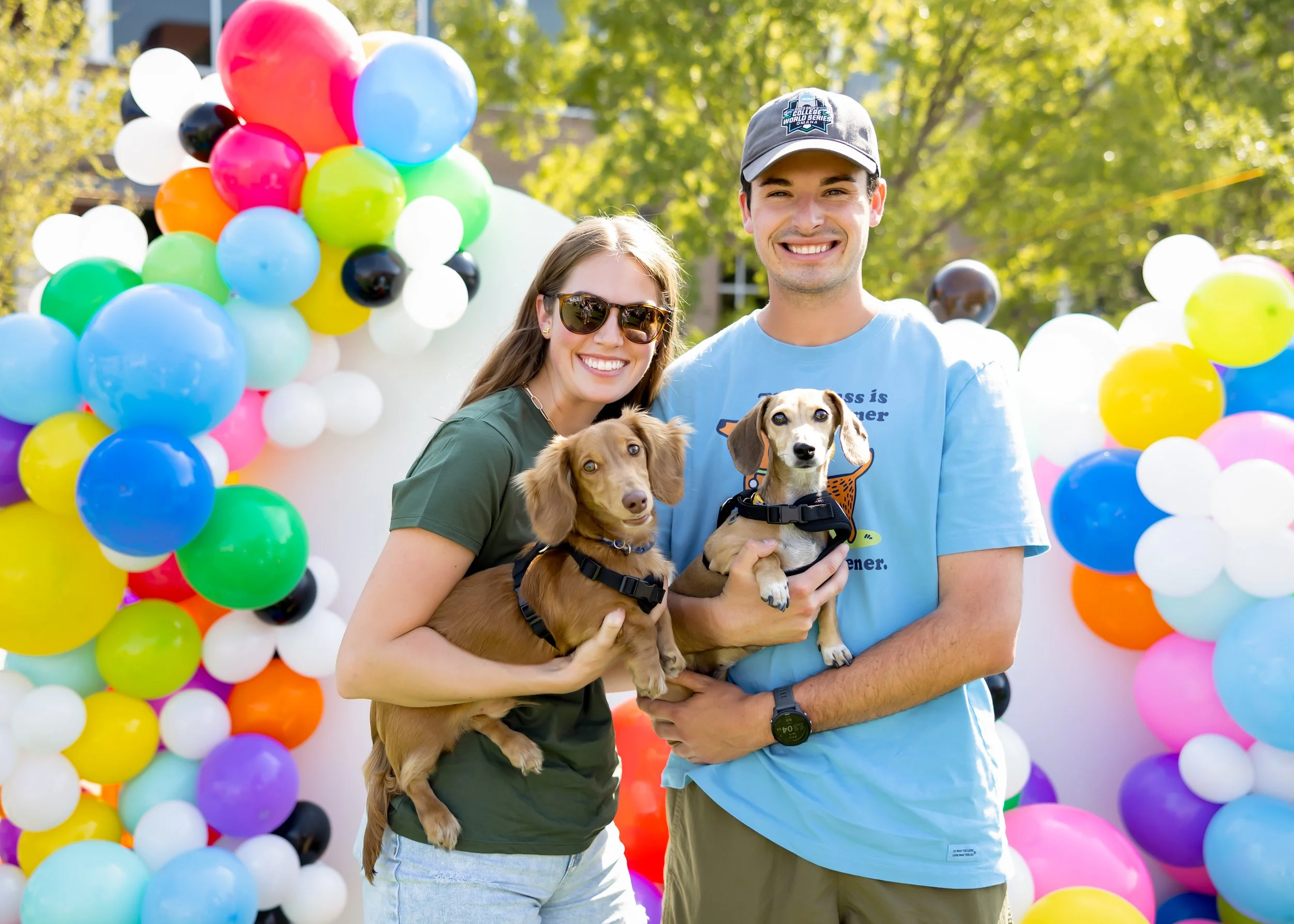 A smiling couple with two Dachshund dogs at a colorful outdoor balloon celebration. The woman is wearing sunglasses and a green shirt, the man is wearing a cap and a light blue shirt. The background shows an arch made of multicolored balloons and green trees.