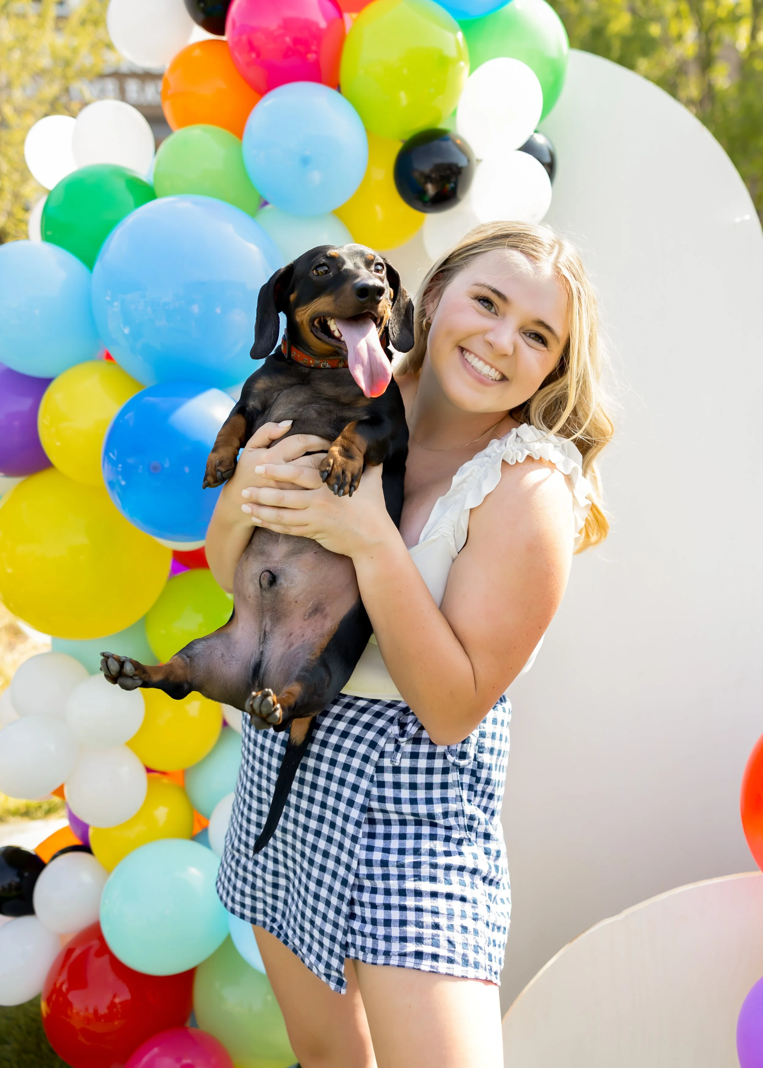 A young woman with blonde hair smiling and holding a happy dachshund puppy in front of a colorful balloon arch outdoors.