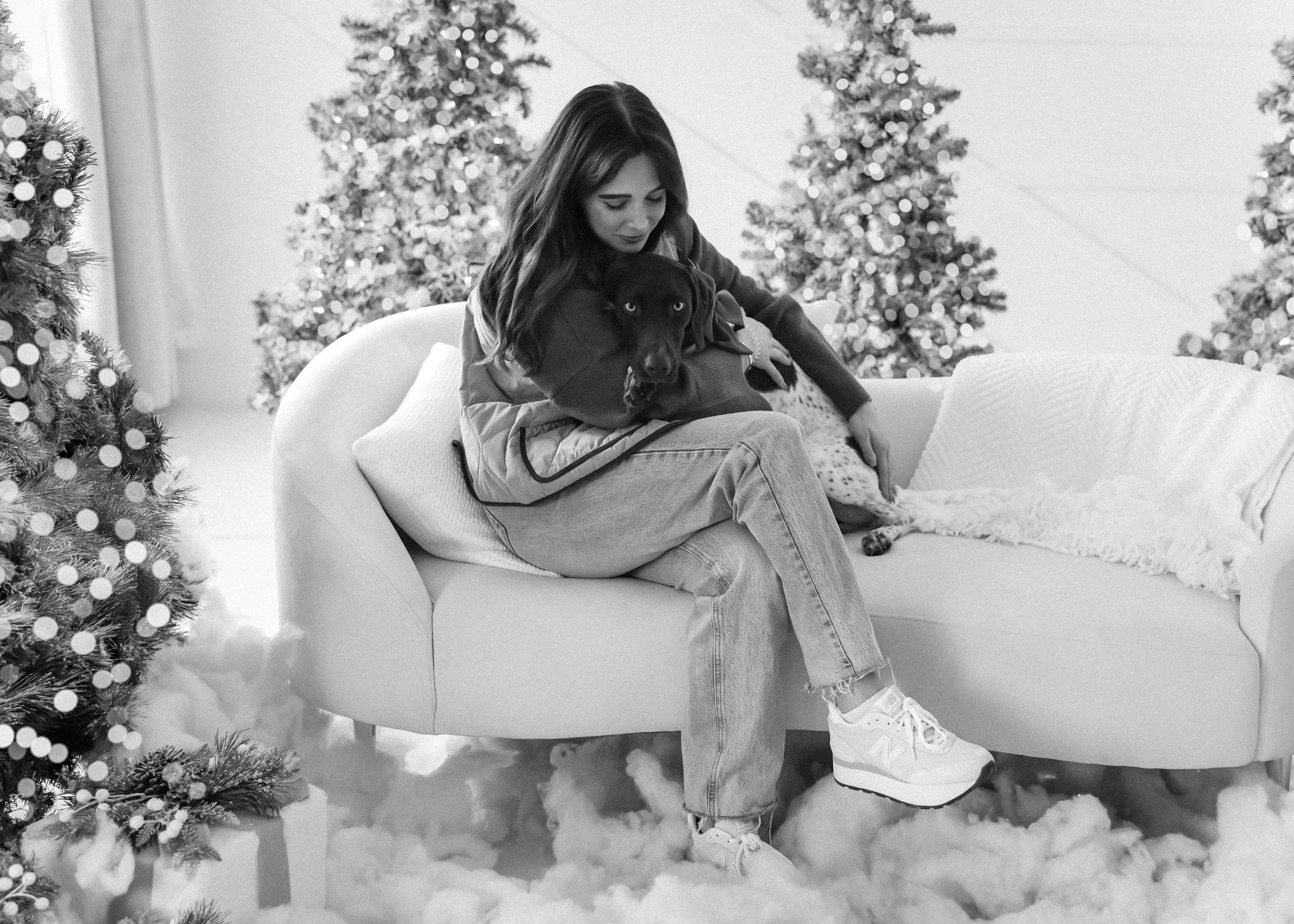 A woman sitting on a white couch holding a black dog in her lap, with Christmas trees in the background and snow on the floor.