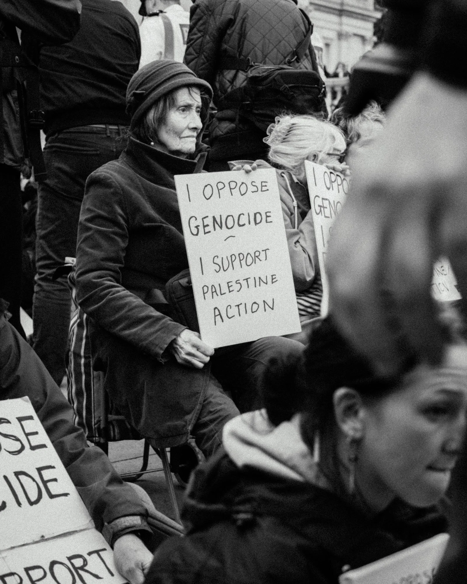 A black and white photo of a woman sitting on a folding chair at a protest, holding a sign that says, 'I oppose genocide – I support Palestine action.' Other protesters with signs are visible around her.
