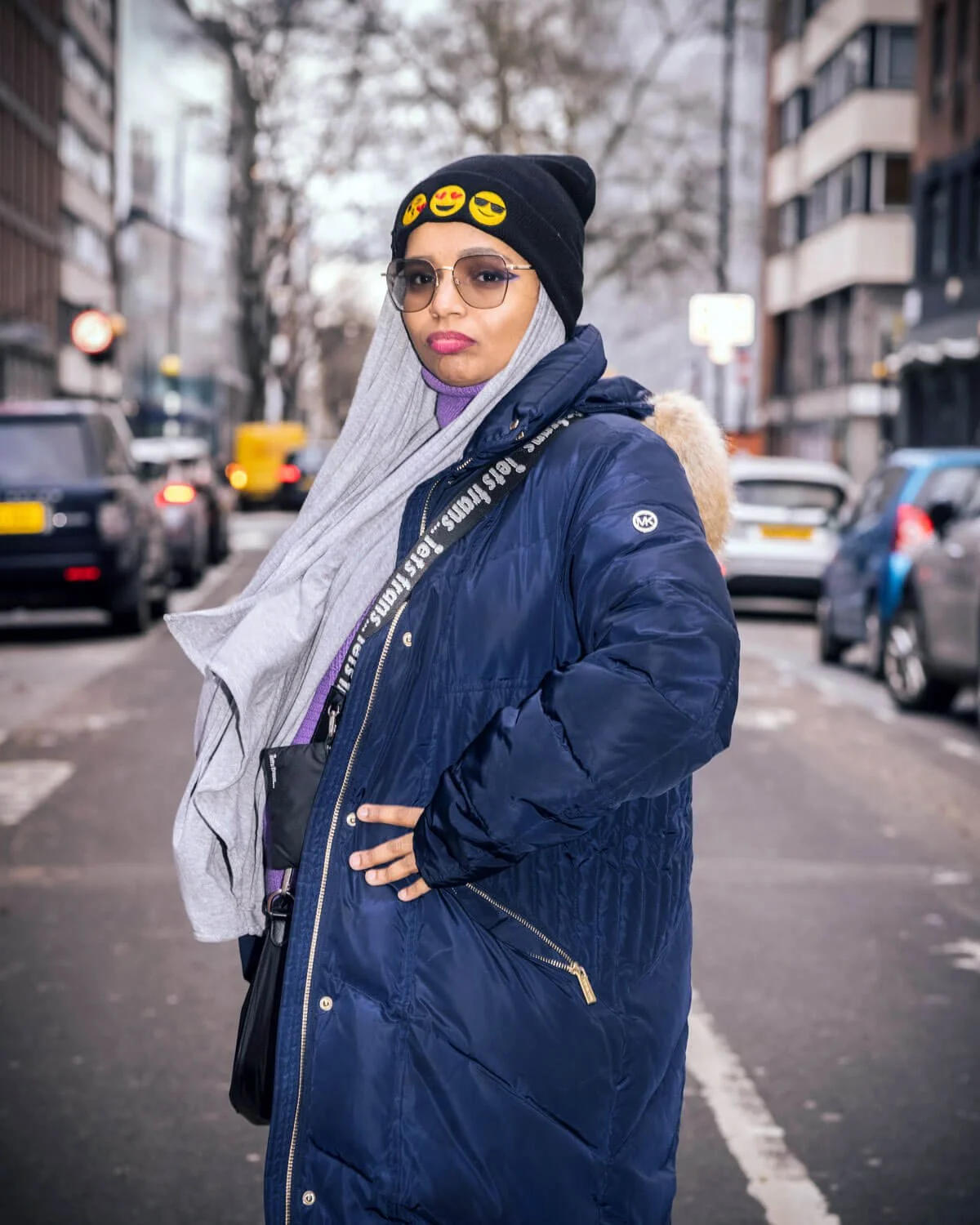 Young woman with blonde hair in a ponytail, wearing a black beanie with emoji patches, sunglasses, a gray hoodie, a purple shirt, and a navy blue puffer coat, standing on a city street with parked cars and leafless trees in the background.