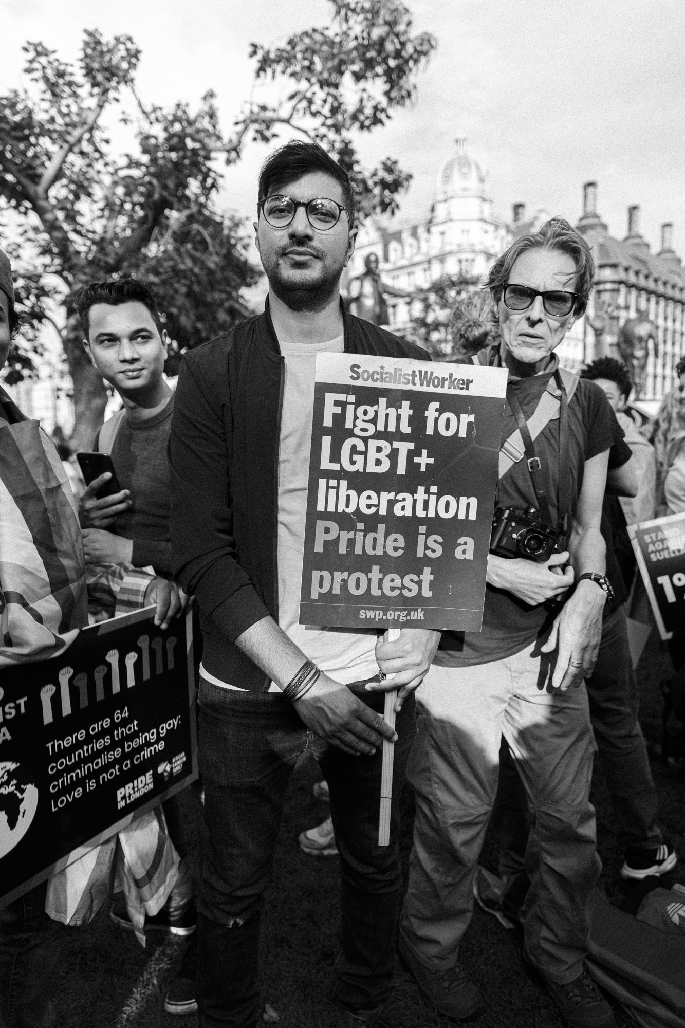 People participating in a pride protest, with one person holding a sign that says, 'Fight for LGBT+ liberation Pride is a protest,' in a city park with historic buildings in the background.