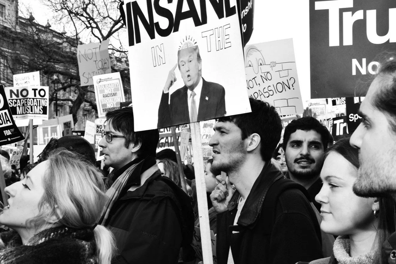 Black and white photo of a diverse crowd protesting, holding various signs including one with an image of Donald Trump. Some signs read "NO TO SCAPEGOATING MUSLIMS," "NOT, NO PASSION?" and other messages.