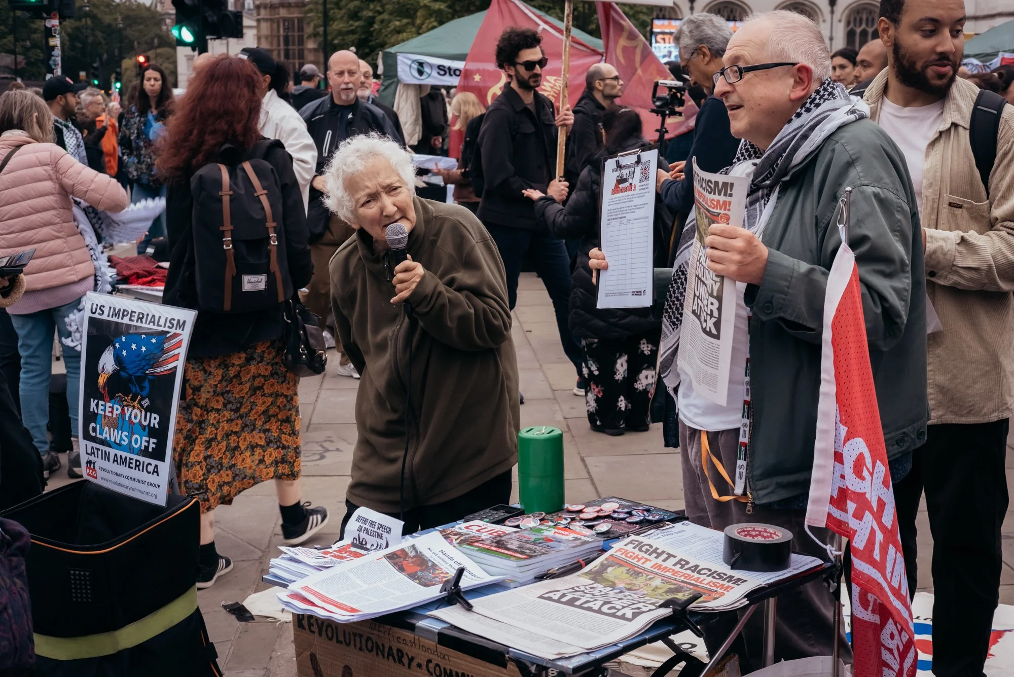 People gathered at a protest or rally, with a table displaying signs, flyers, and buttons promoting anti-racism and anti-imperialism messages. An elderly woman is speaking into a microphone, and others are holding signs and engaging in discussion.