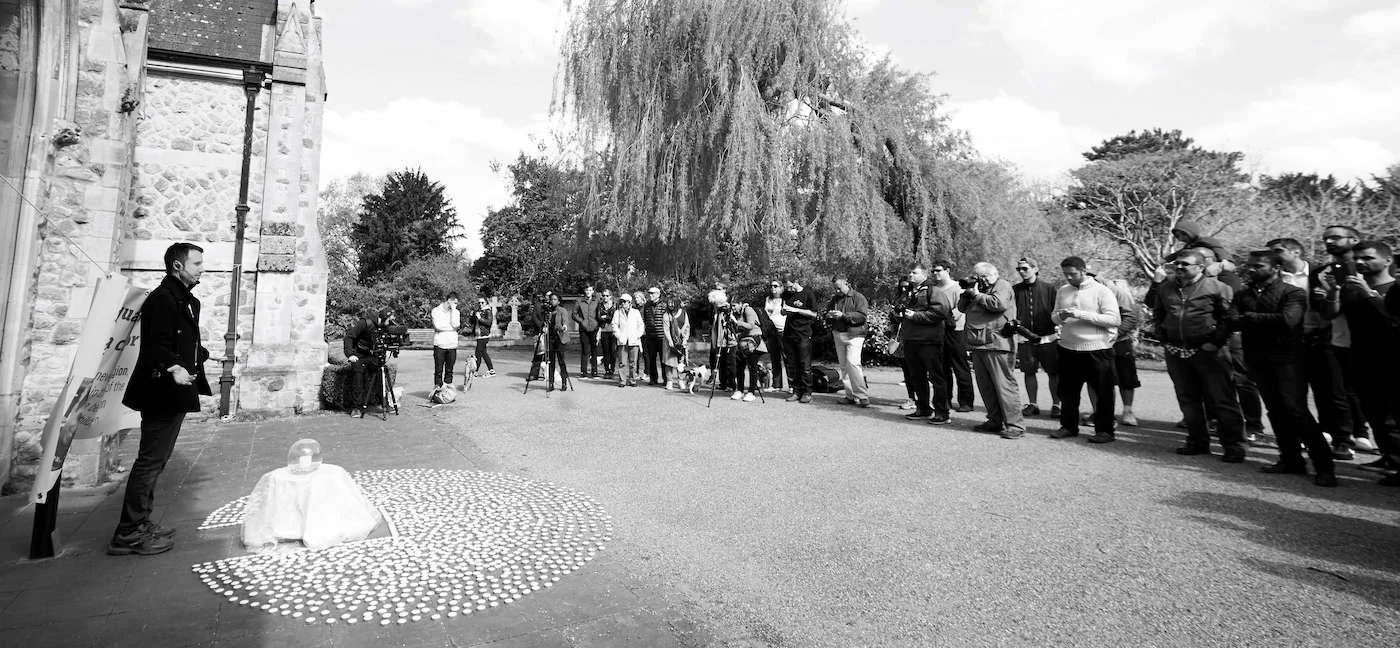 A man stands with his hands in his pockets facing a group of people gathered outdoors, some holding cameras. There is a small table with a white cloth and candles arranged in a circle on the ground in front of him, near an old stone building. The set