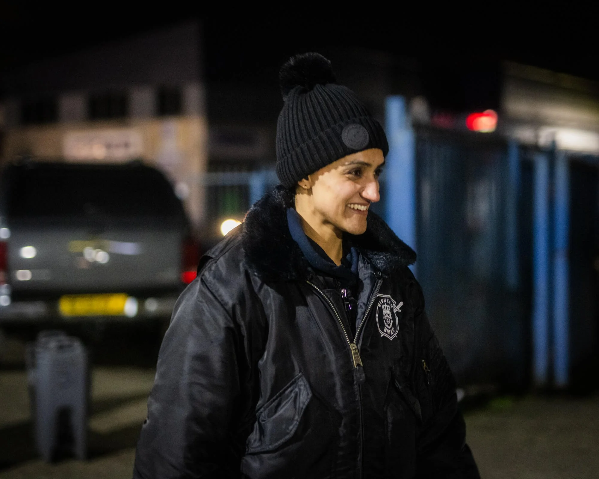 A woman in police uniform smiling outdoors at night, wearing a black beanie and jacket.