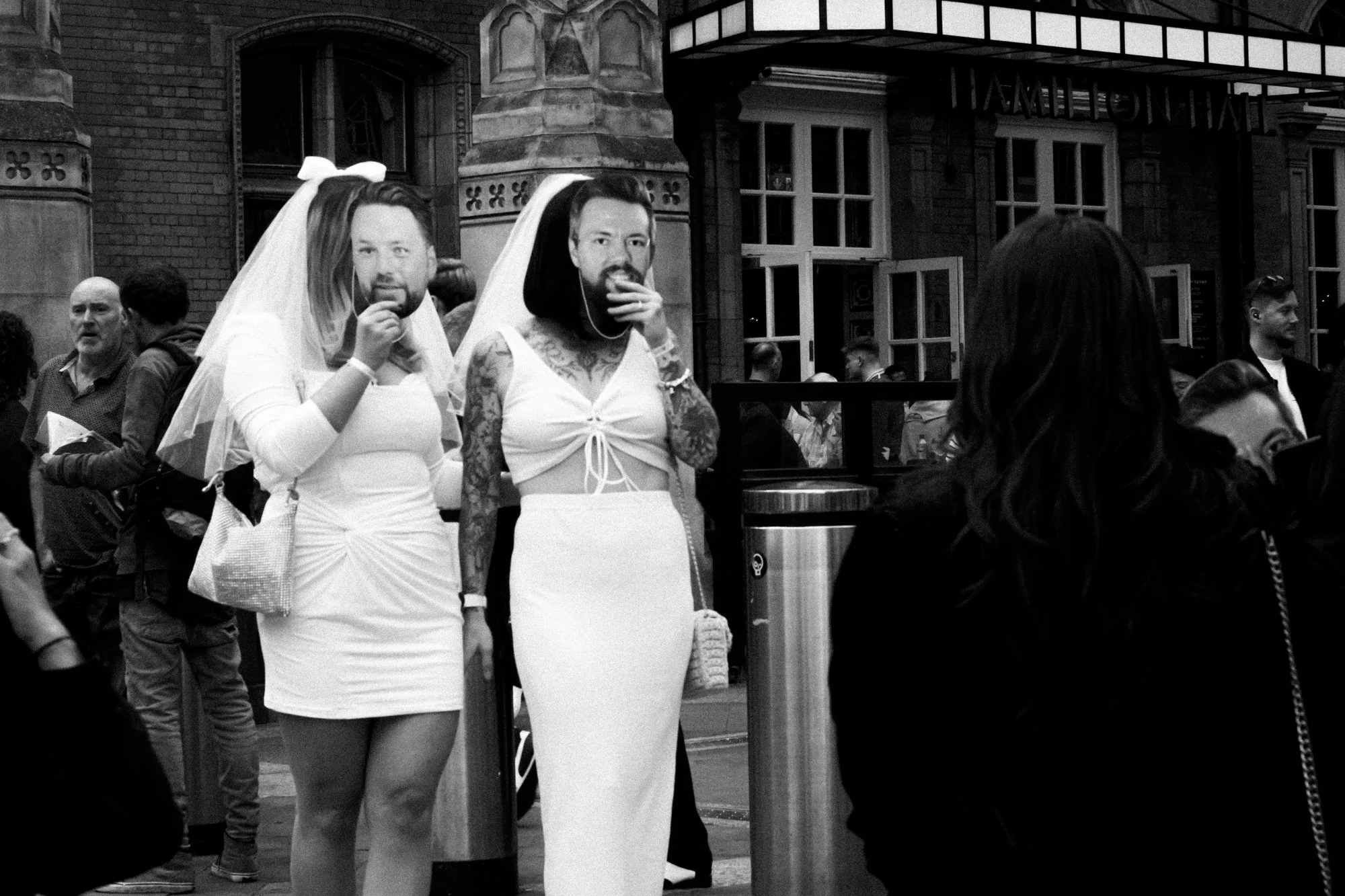 Two women dressed as brides with veil and wedding dress, standing among a crowd outside a building, one wearing a white dress with a veil, and the other with tattoos wearing a white crop top and long skirt, facing the camera.
