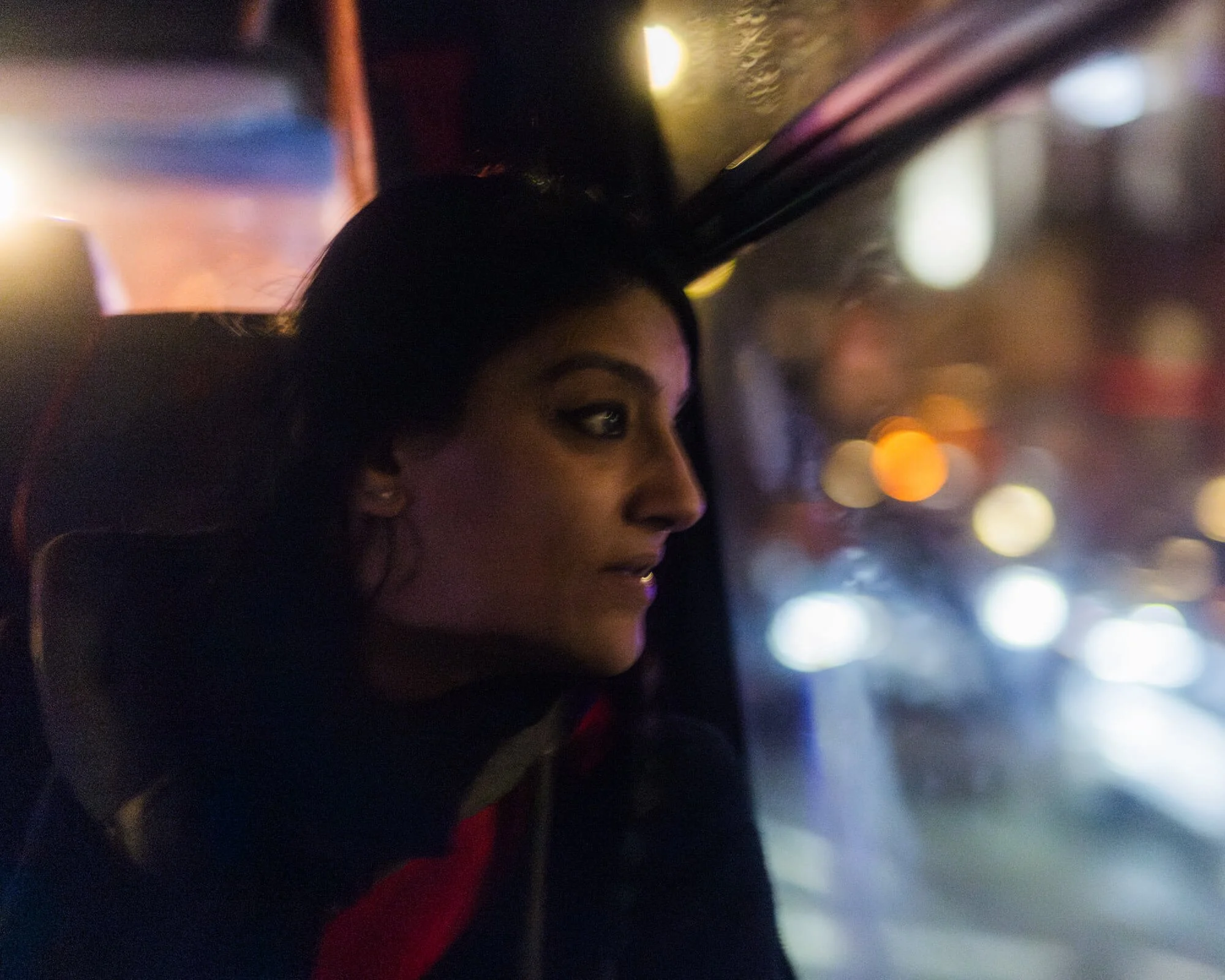 A woman with dark hair and makeup sitting inside a vehicle at night, looking out the window with city lights in the blurred background.