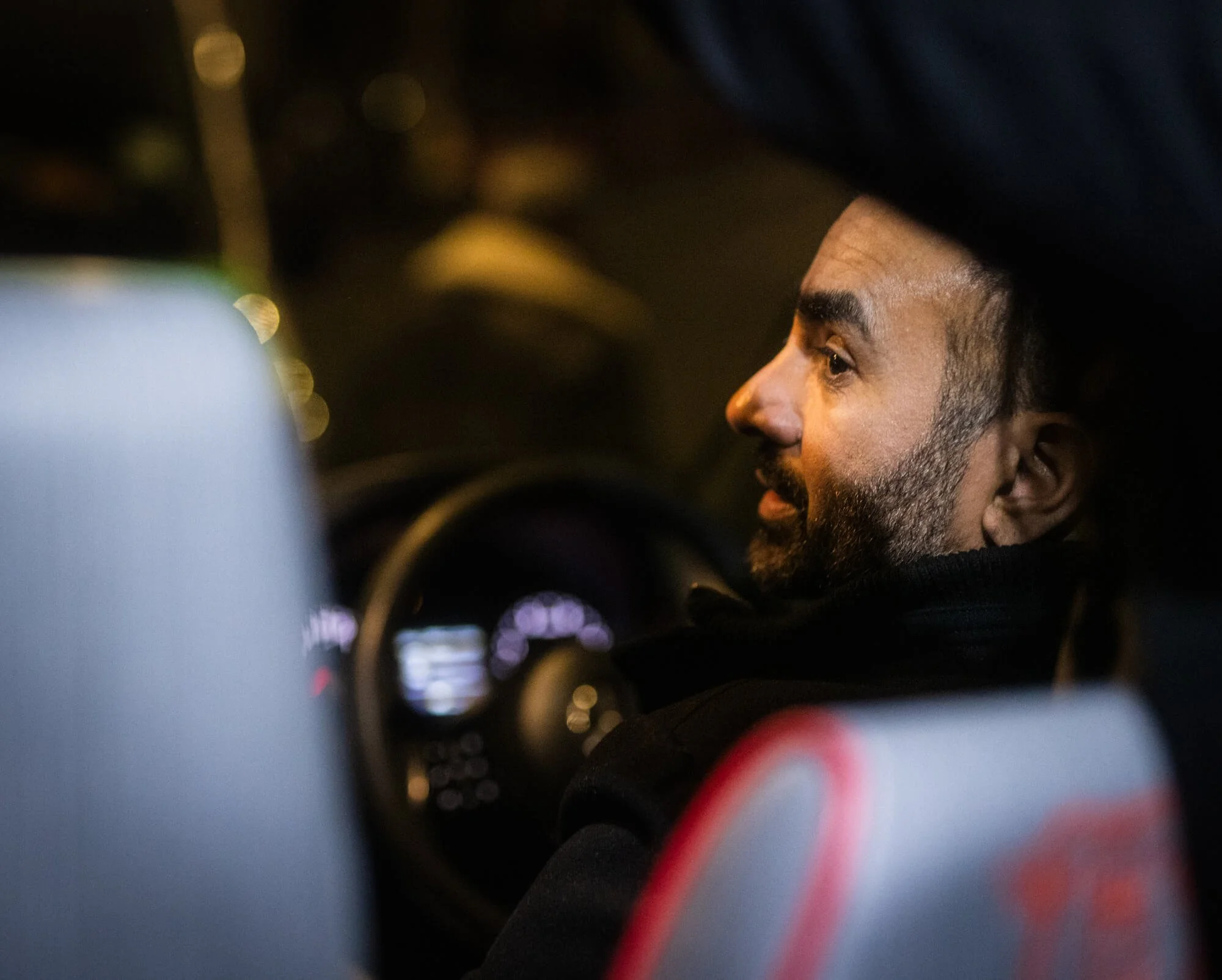Side profile of a man with a beard and dark hair sitting in a car at night, illuminated by dashboard lights.
