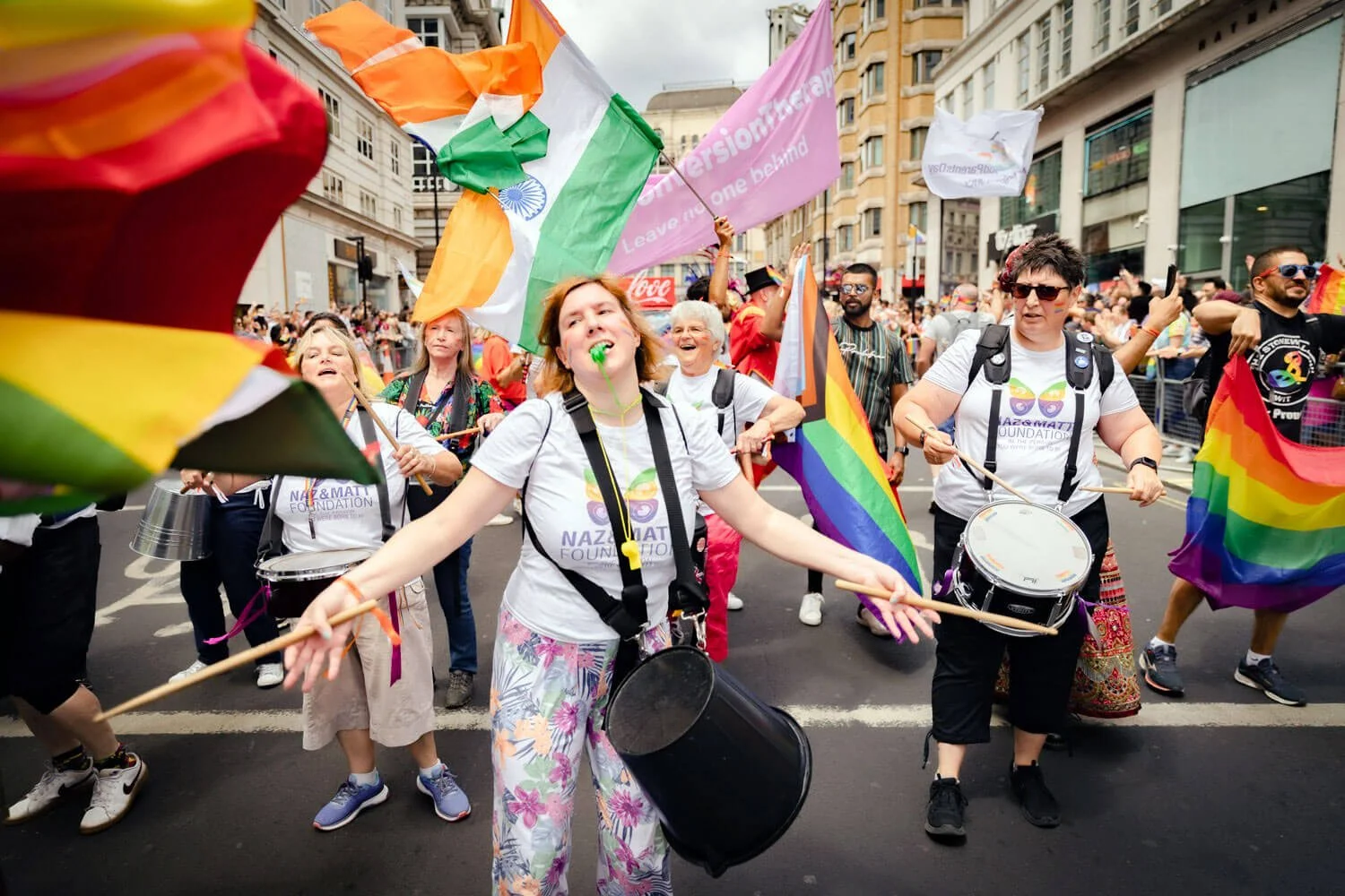 People participating in a pride parade, holding rainbow flags and placards, some with flags of India and LGBTQ+ symbols, marching down city streets with buildings in the background.