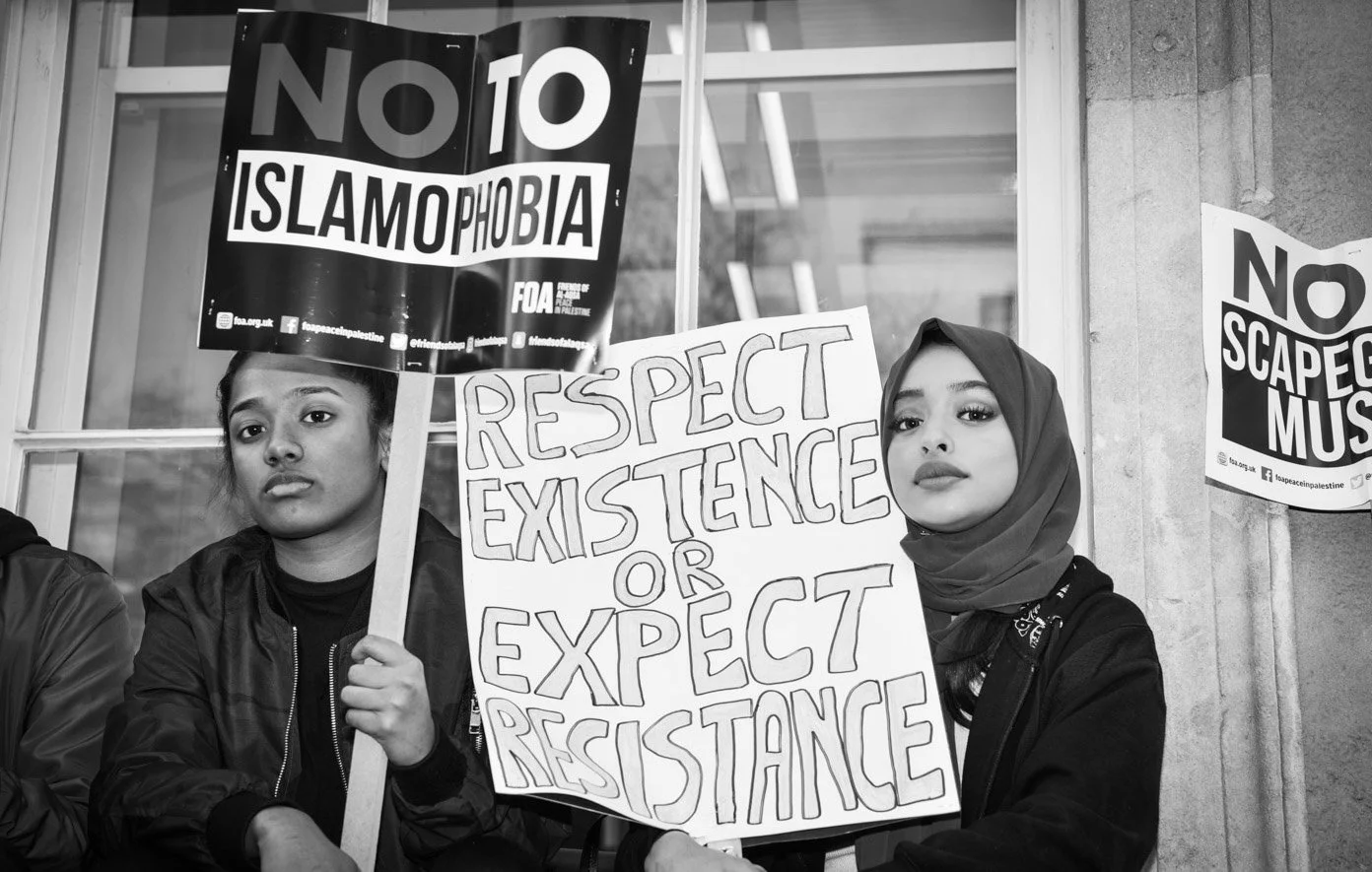 Two young women participating in a protest or demonstration, holding signs with messages such as "NO ISLAMOPHOBIA" and "RESPECT EXISTENCE OR EXPECT RESISTANCE," standing in front of a building with glass windows.