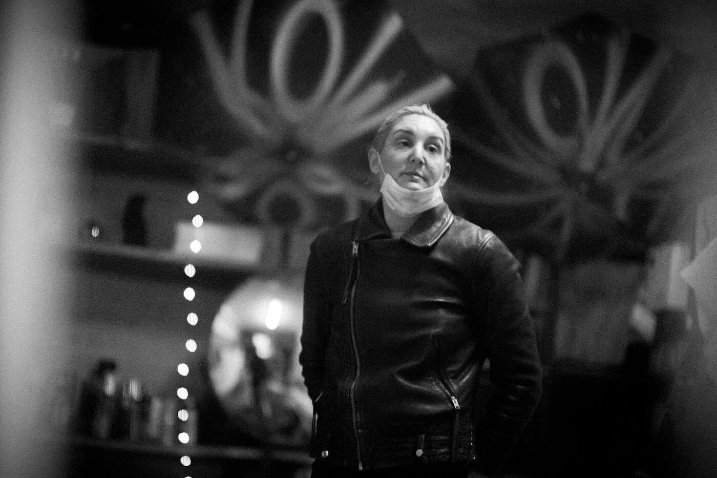 A woman with a face mask pulled down to her chin, wearing a leather jacket, standing in front of a mirror with a decorative design, in a dimly lit space.