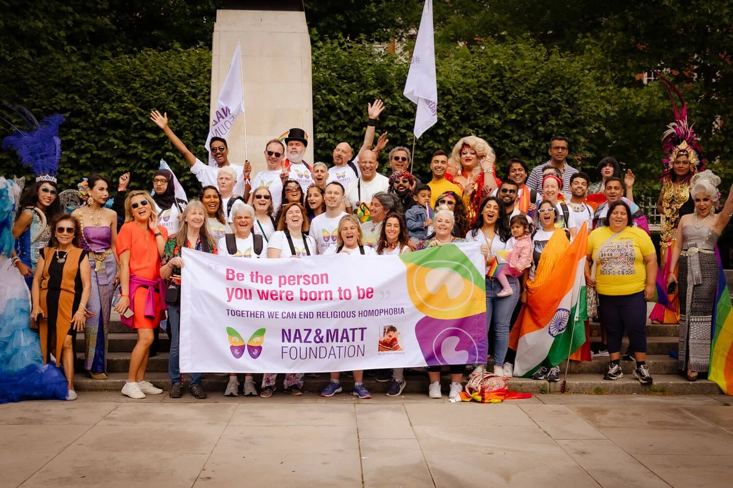 A diverse group of people holding a large banner and flags during a pride event, celebrating LGBTQ+ acceptance and supporting the Naz & Matt Foundation against religious homophobia. The group is smiling and dressed brightly, with some in costumes and others in casual attire, standing on steps in an outdoor park setting.