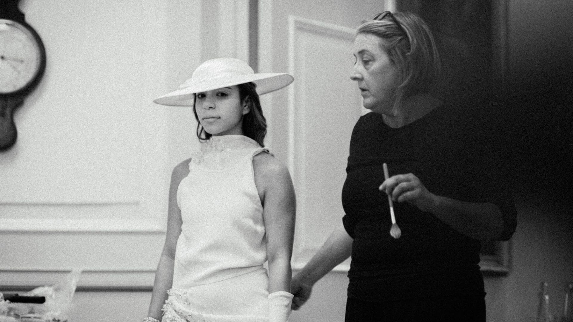 A young girl wearing a wide-brimmed hat and a sleeveless dress receiving makeup or face paint from an adult woman holding a brush in a domestic setting.