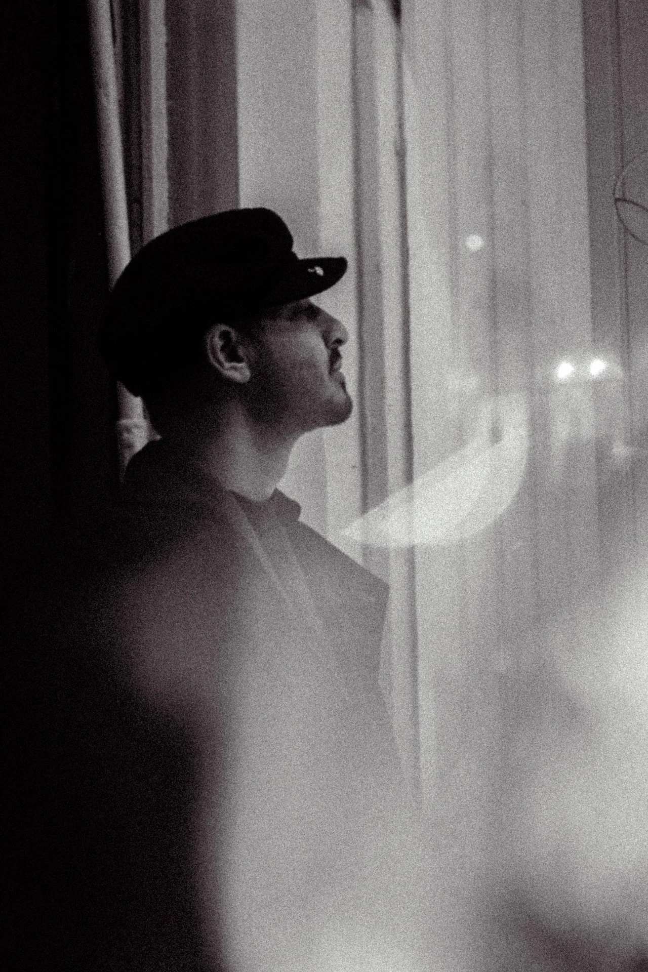 A person in a dark cap looking out a window at night, visible in profile with a contemplative expression, through glass that reflects some city lights.