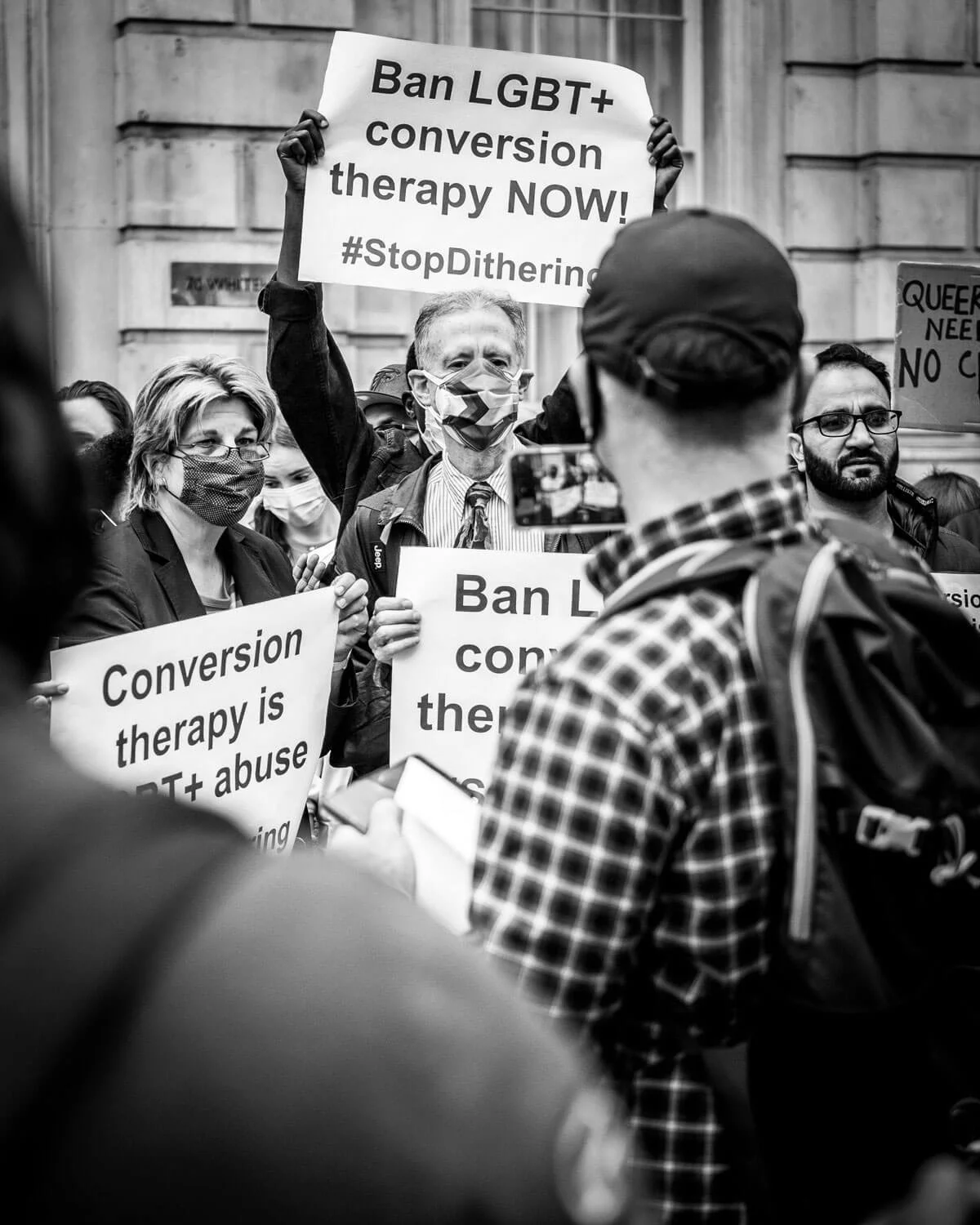 A black and white photo of a protest with people holding signs advocating against LGBT+ conversion therapy, with the woman in the foreground wearing a checkered shirt and backpack, and others wearing masks.
