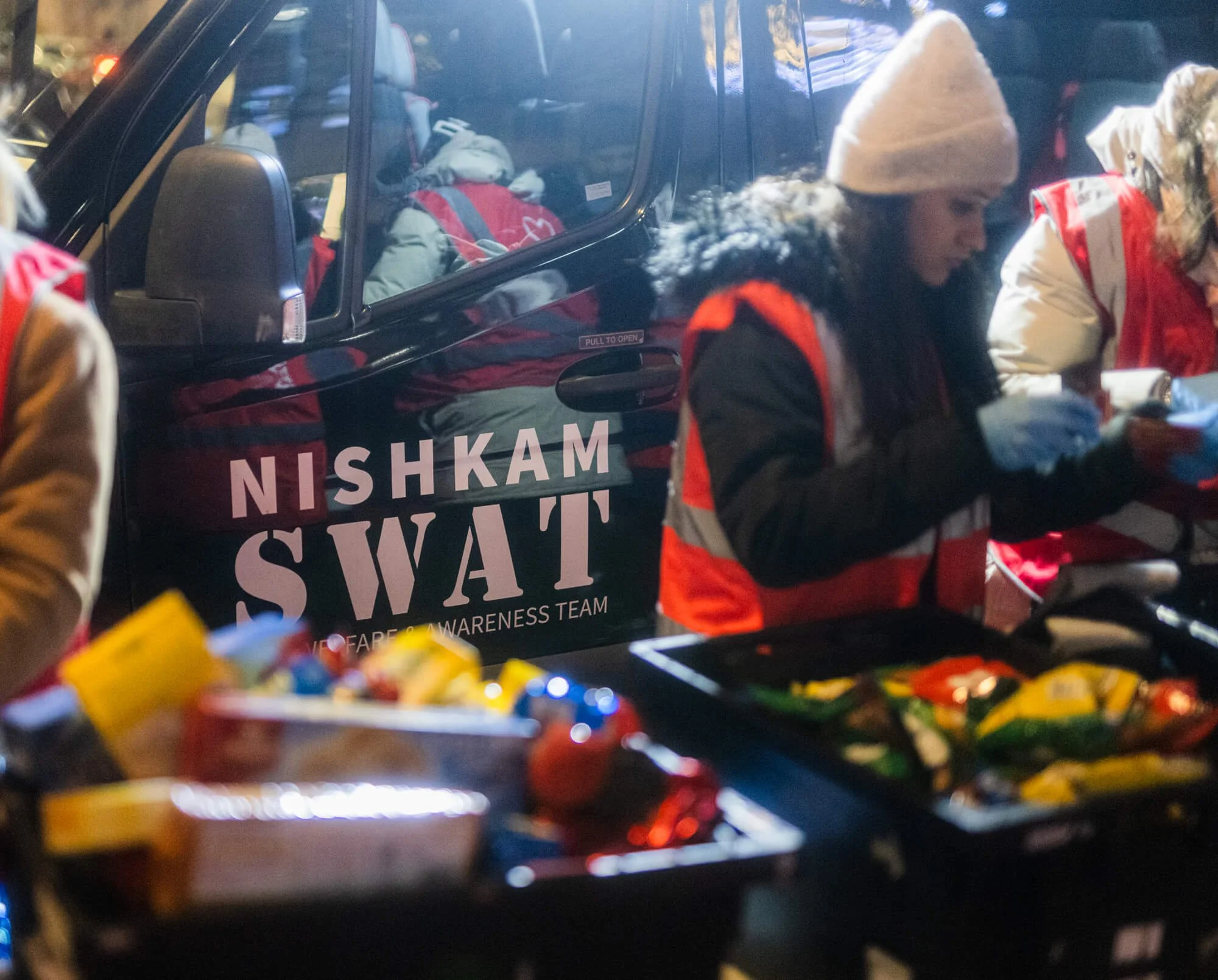 Volunteers from the Nishkam SWAT team preparing food at a community event, with a vehicle in the background bearing their team name.