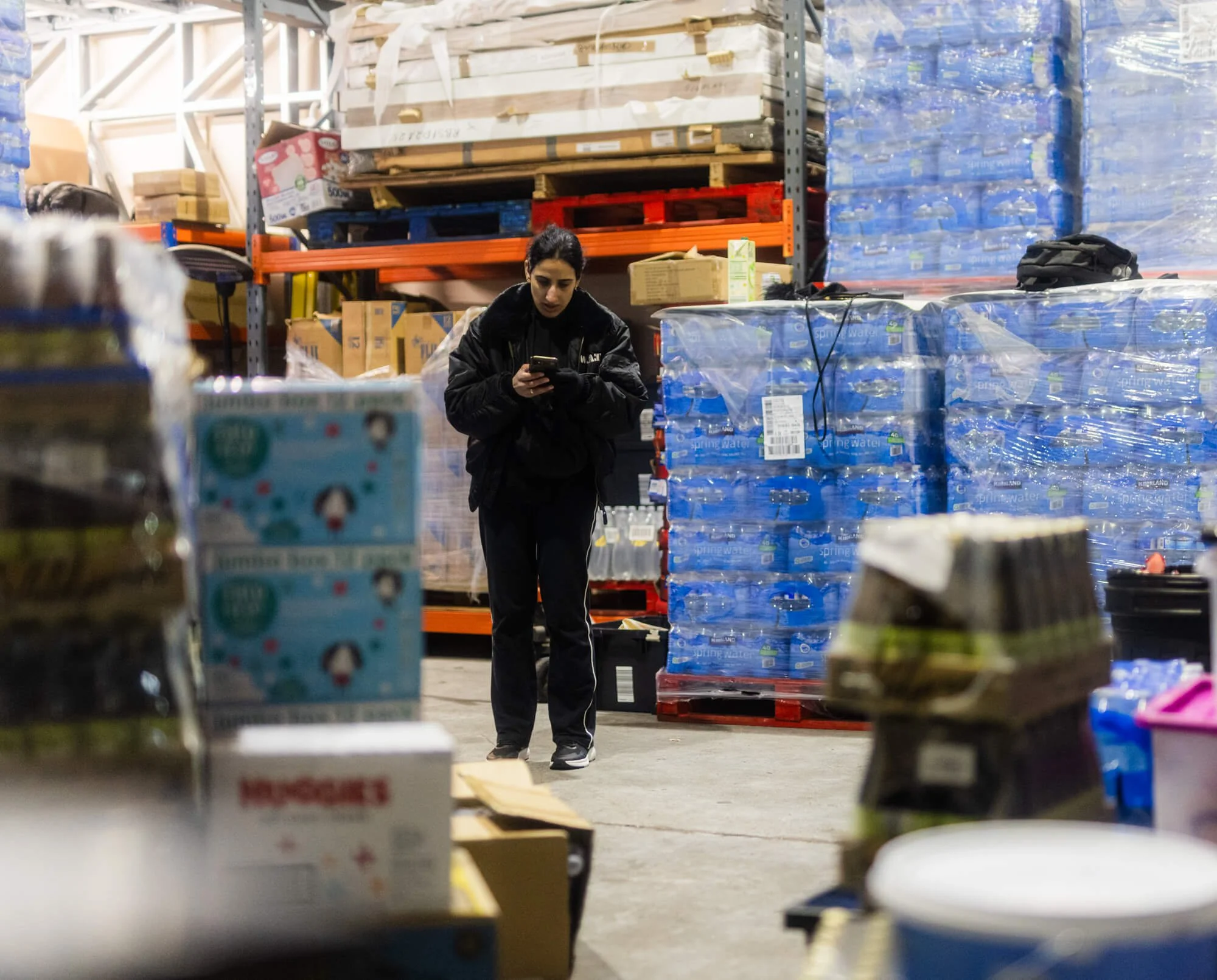 A woman in black clothing standing in a warehouse aisle, looking at her phone surrounded by shelves of bottled water and cardboard boxes.