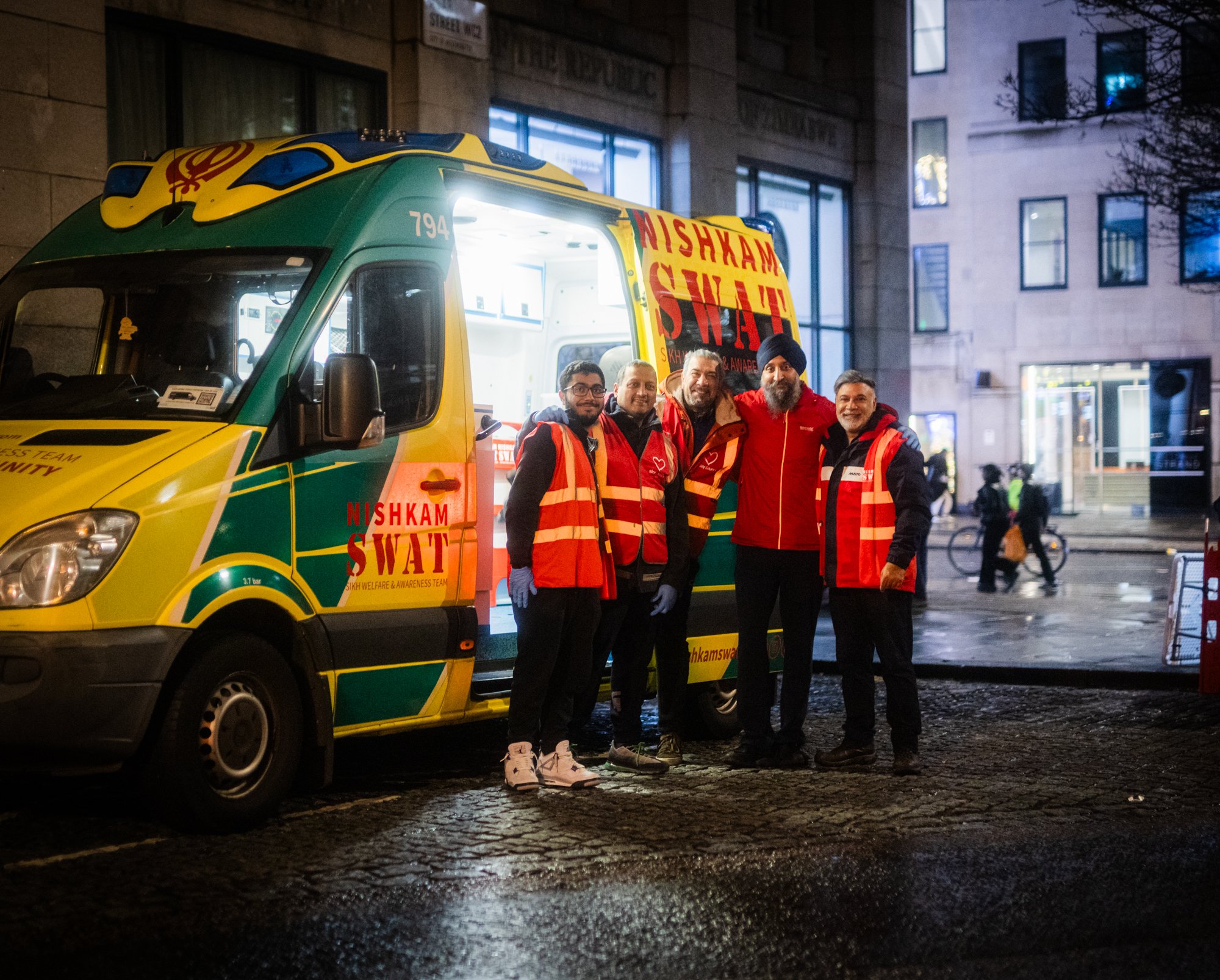 Five people standing in front of a green and yellow Nishkam SWAT van at night, wearing red and black uniforms with reflective vests, in an urban environment with buildings and pedestrians in the background.