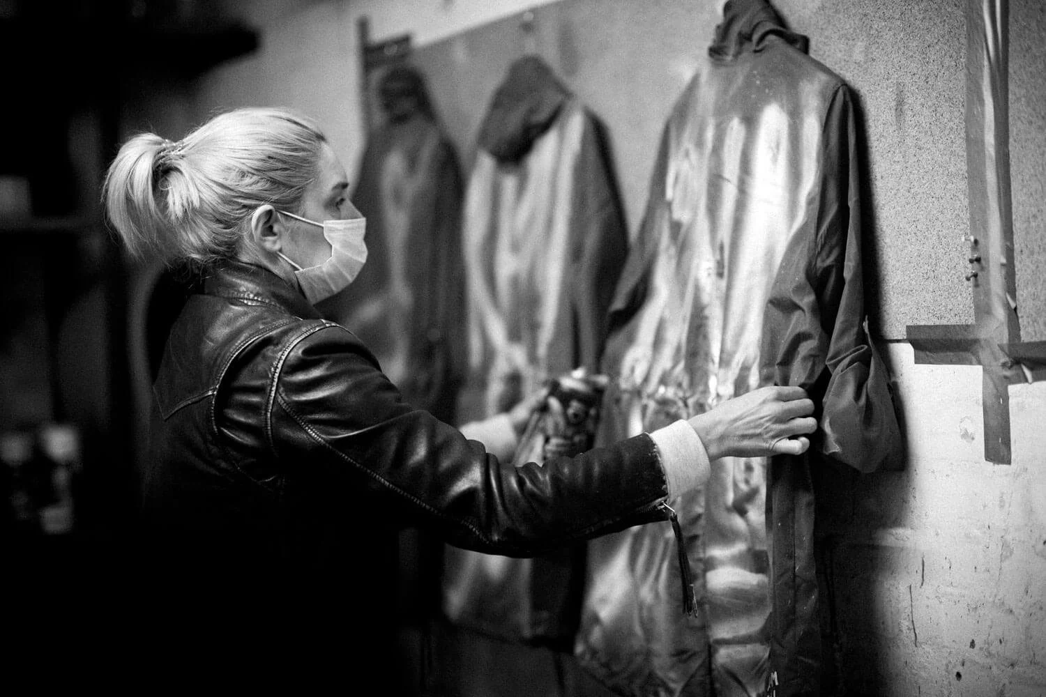 A woman wearing a face mask and a leather jacket looks at a row of jackets or costumes hanging on a wall, touching one of them.