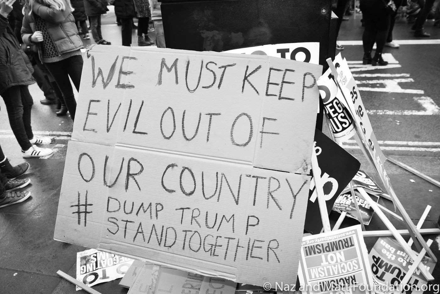 A protest sign reading "We must keep evil out of our country # Dump Trump" surrounded by other protest signs on a city street.