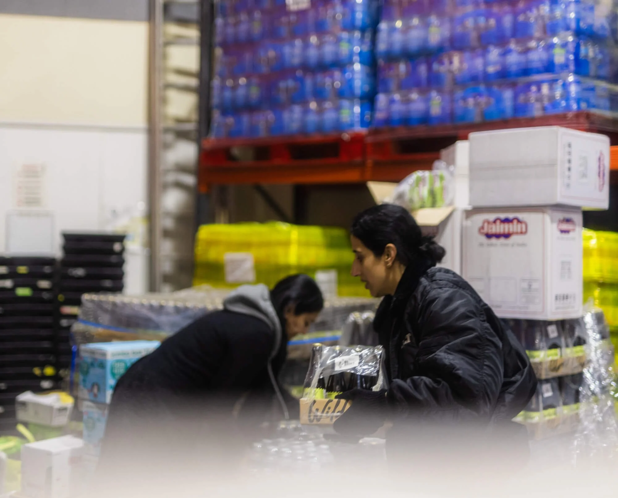 Two women working in a warehouse surrounded by stacked bottled water and boxes.