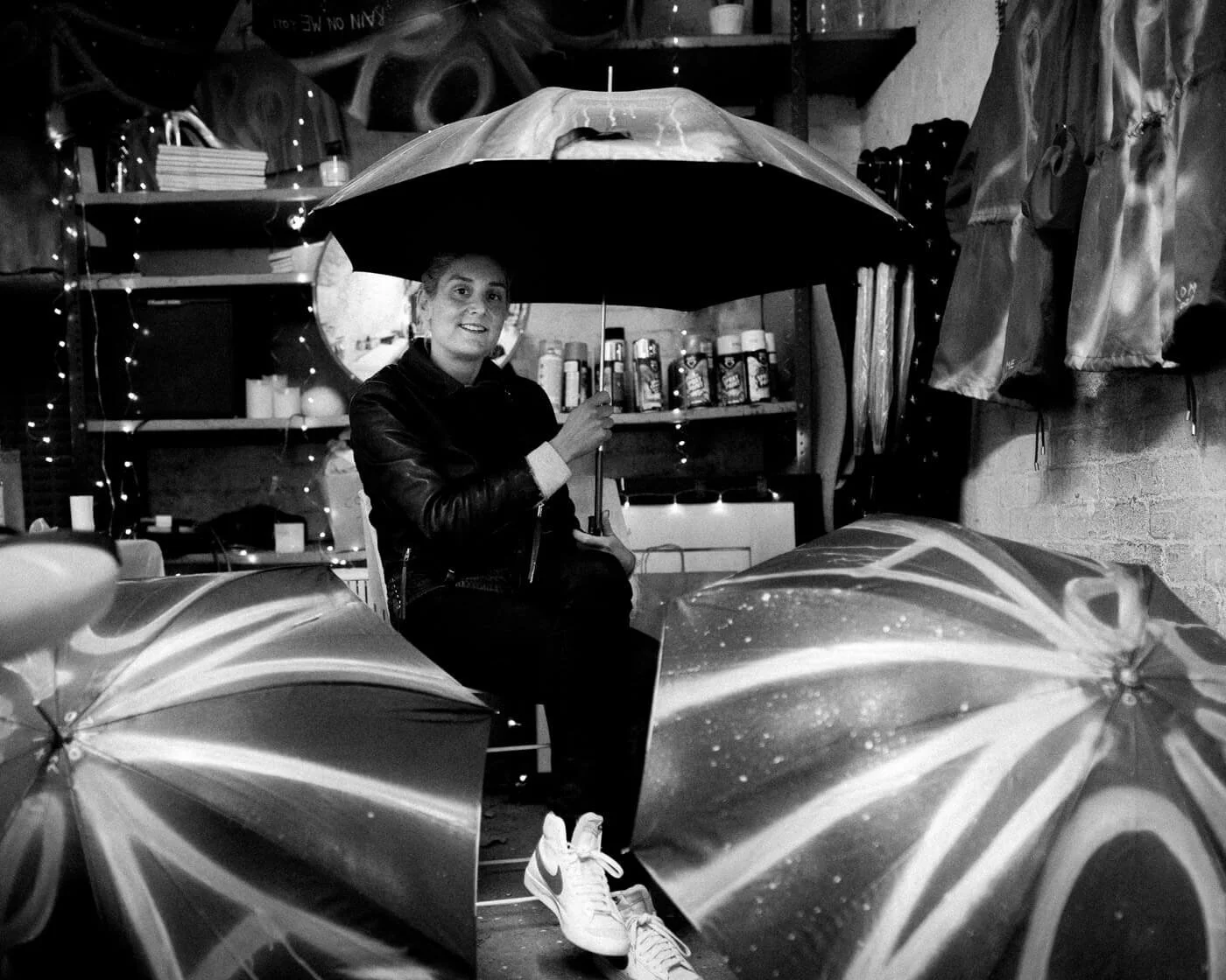 A woman sitting on a chair holding an umbrella, surrounded by other umbrellas, inside a room with shelves and hanging decorations in black-and-white.