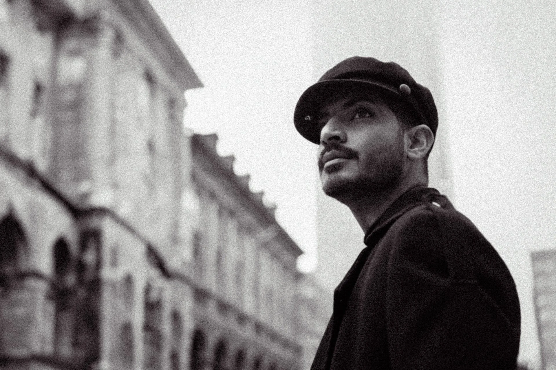 A young man with a beard wearing a black hat and coat, looking upwards with a cityscape in the background.