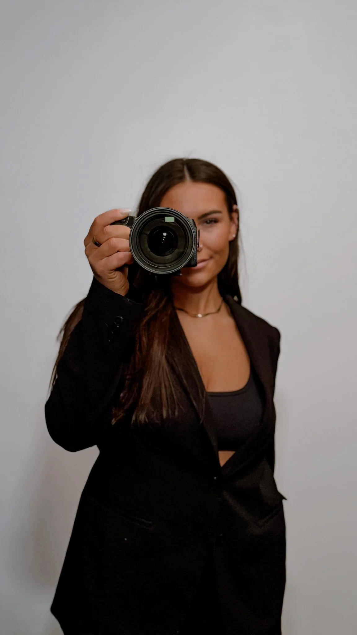 Woman in black blazer and top holding a camera in front of a white wall.