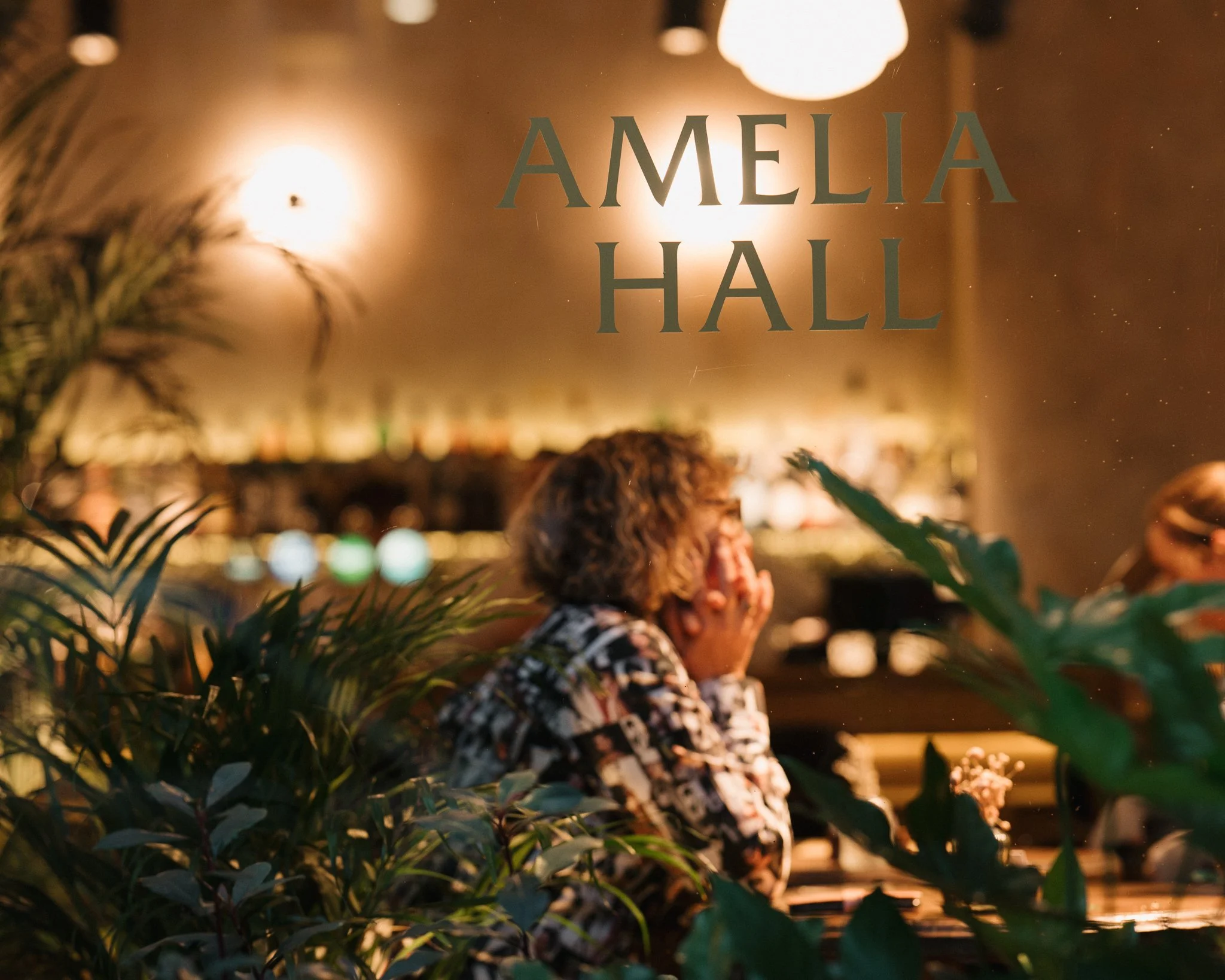 A woman with curly hair plays the piano in a softly lit room behind a glass door labeled 'Amelia Hall.' She covers her mouth with one hand, appearing emotional, with green plants in the foreground and a blurred background of warm lighting.