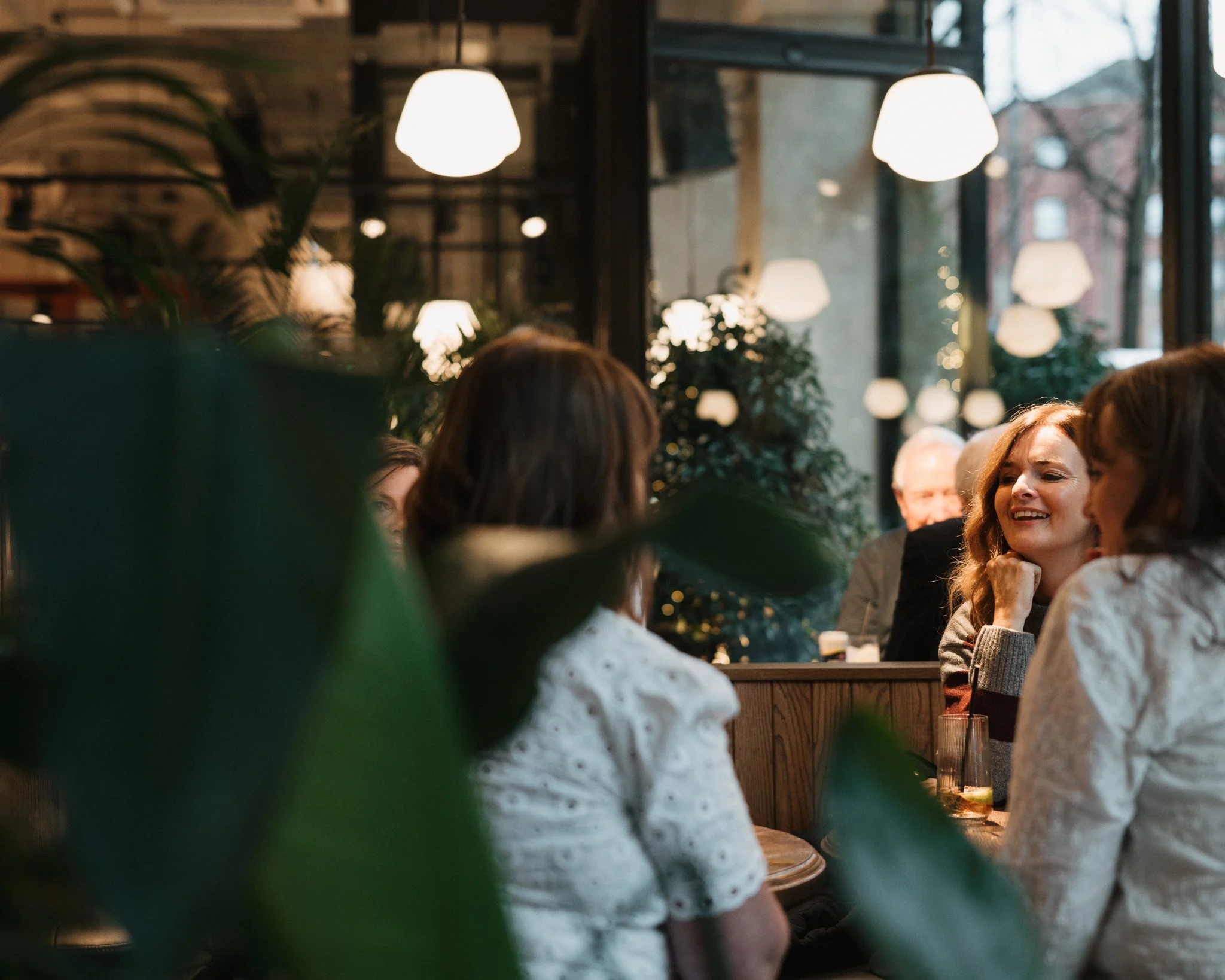 People sitting at a table in a cozy, warmly lit restaurant enjoying conversation and drinks, with large windows and hanging lights.
