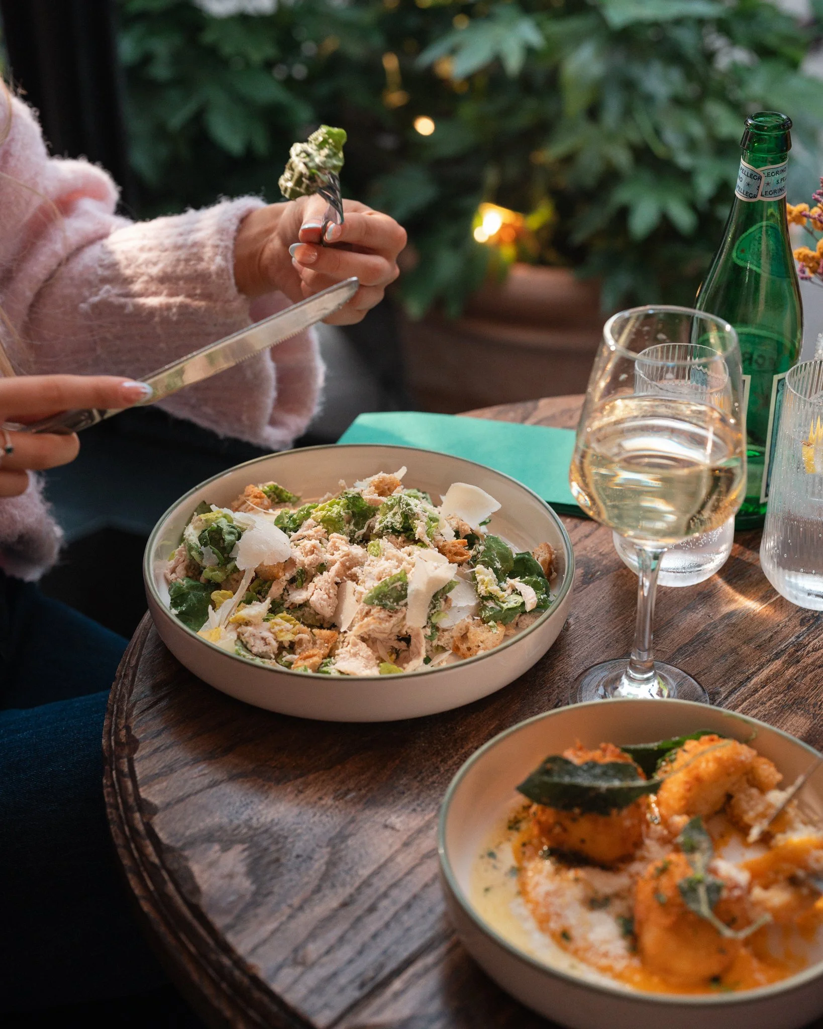 A person is seen eating a salad at a wooden table set with glasses of water and wine, a green glass bottle, and bowls of food; leafy plants are in the background.